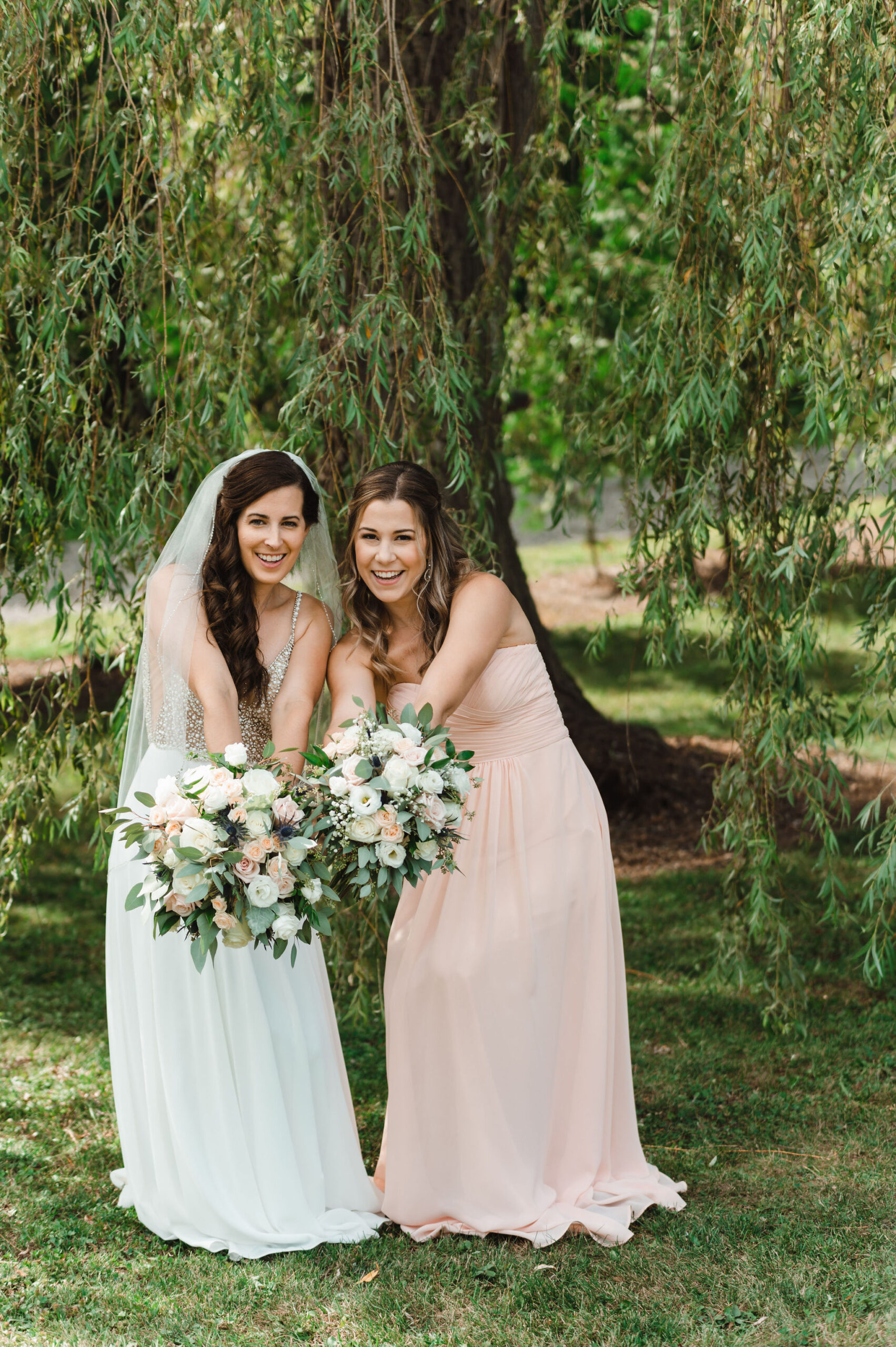 bride and her bridesmaid holding out floral bouquets underneath a willow tree during her special wedding at Carvers cottage in Pickering