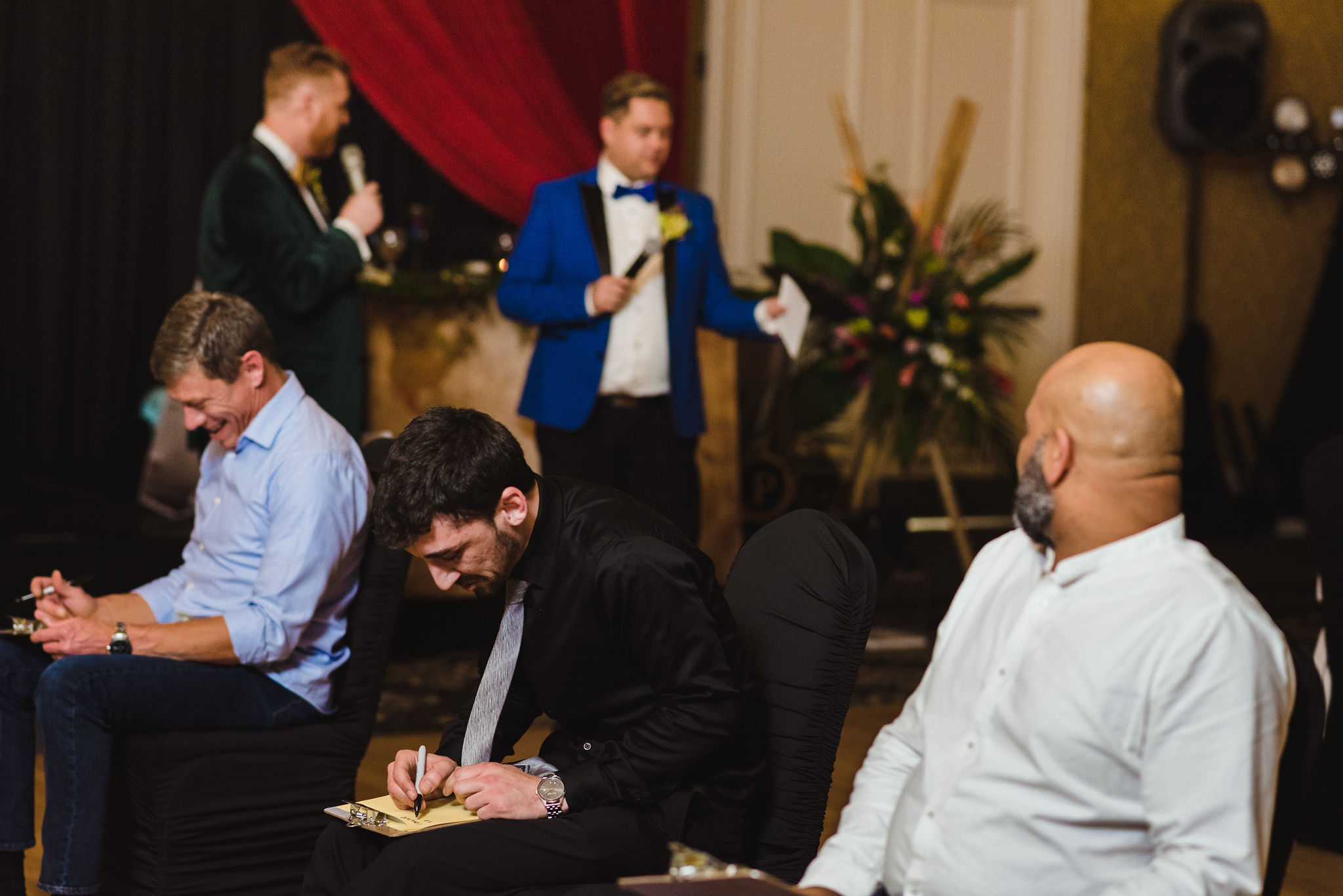 grooms reading game instructions into their microphones while seated guests are scribbling away on clipboards during a fun wedding at the Hilton Fallsview in Niagara Falls grooms reading game instructions into their microphones while seated guests are scribbling away on clipboards during a fun wedding at the Hilton Fallsview in Niagara Falls