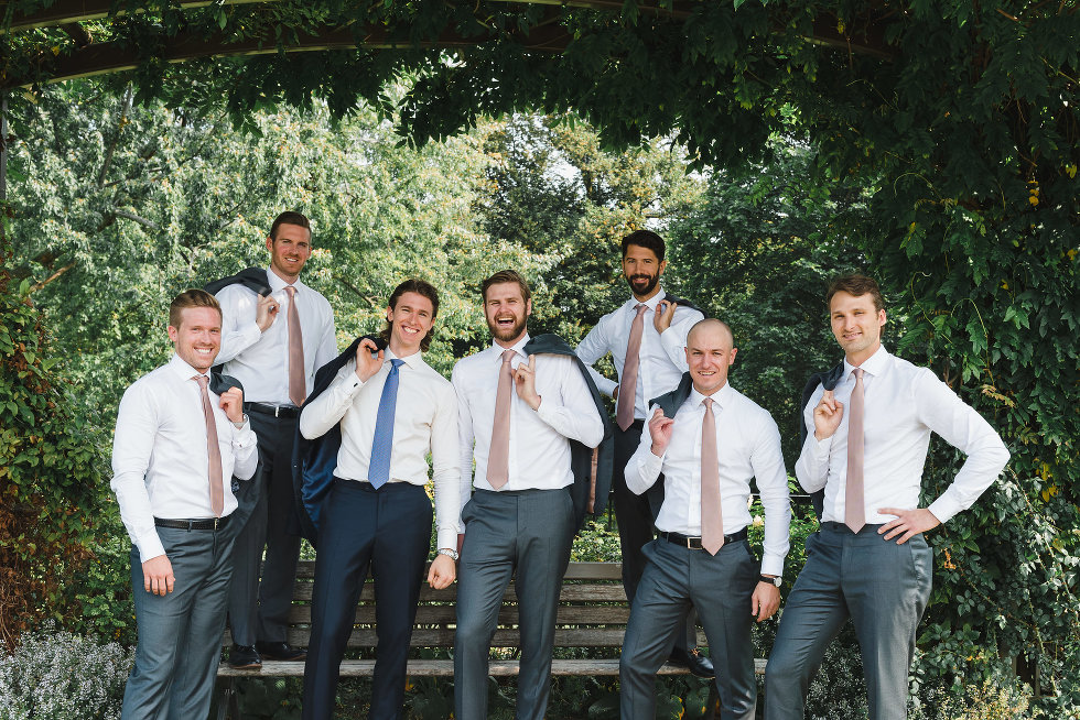 groom and his groomsmen with jackets draped over shoulders as they stand around park bench Toronto wedding photography