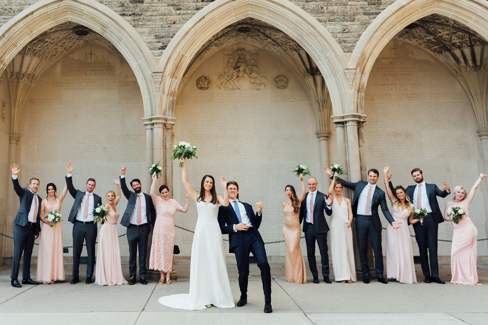 bride, groom, and wedding party with hands in the air in front of church arches Toronto wedding photography