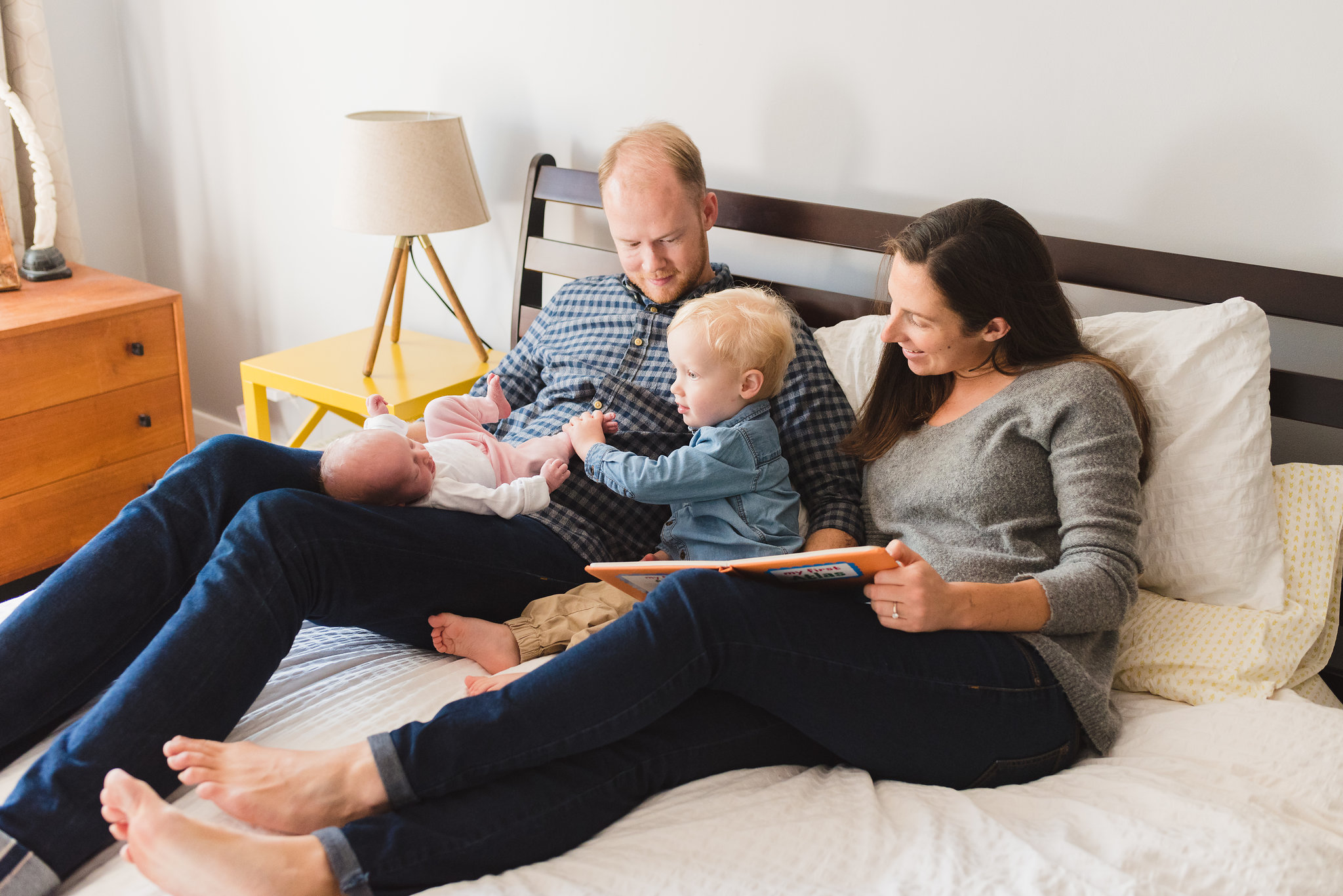 parents lying on bed with baby in fathers lap and toddler in the middle while mother reads a book Toronto photographer Gillian Foster