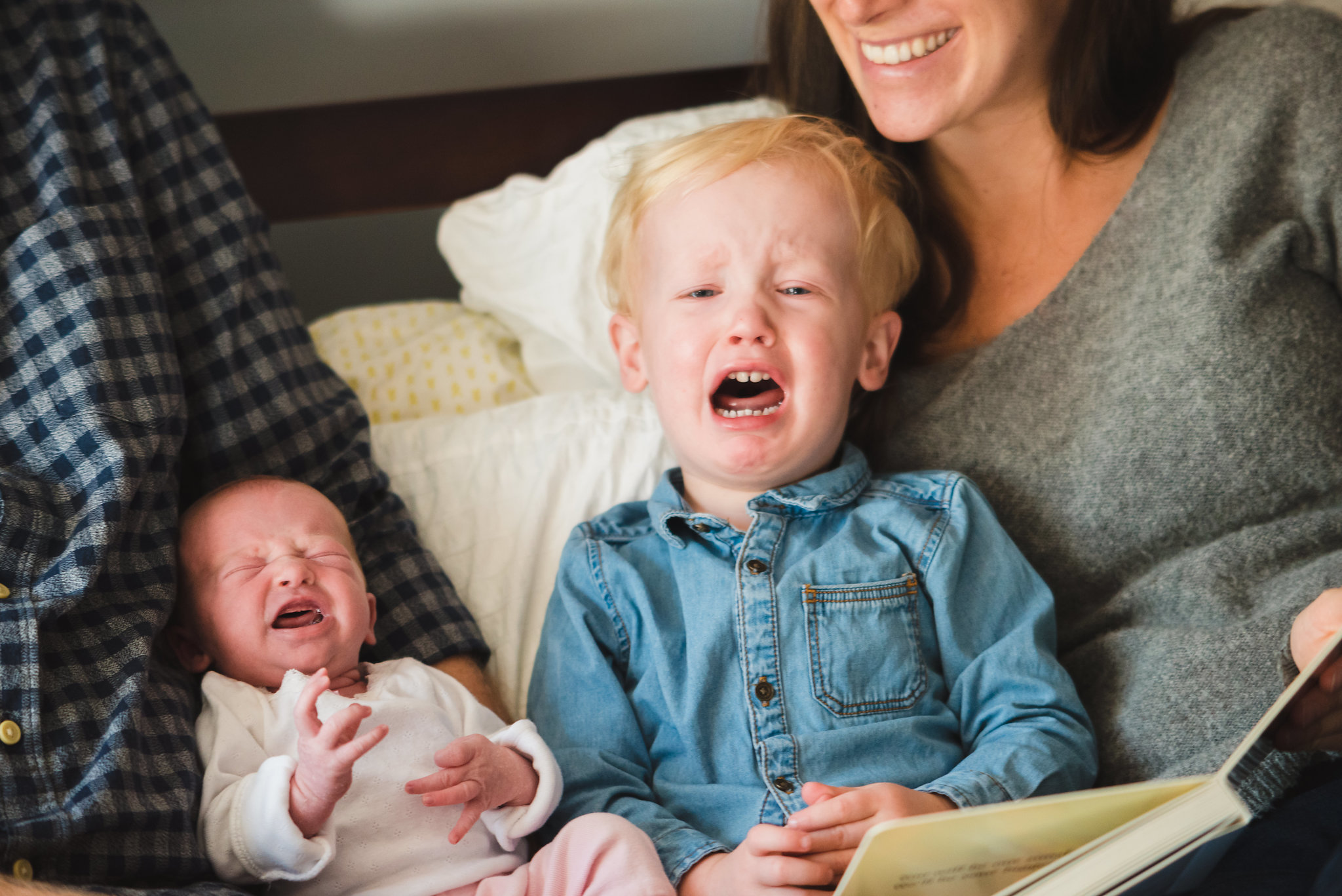 parents lying in bed with crying newborn and crying toddler while mother reads book to toddler Toronto