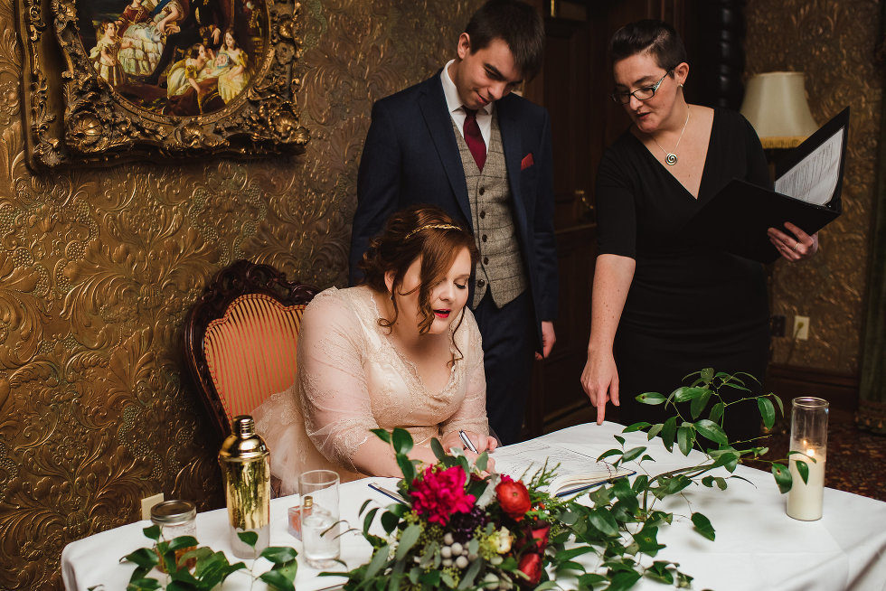 bride seated as she signs wedding certificate with groom and officiant looking on Niagara wedding photography 