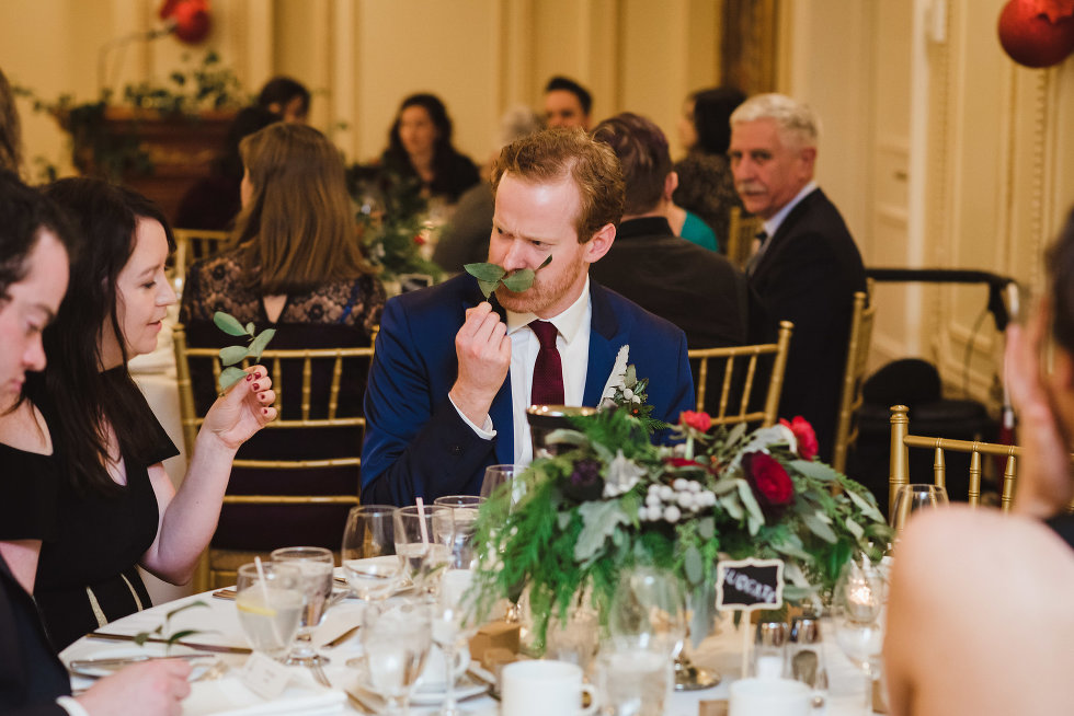 groomsman sitting down at table holds green leaves up to his lips to make a fake moustache Niagara wedding photography