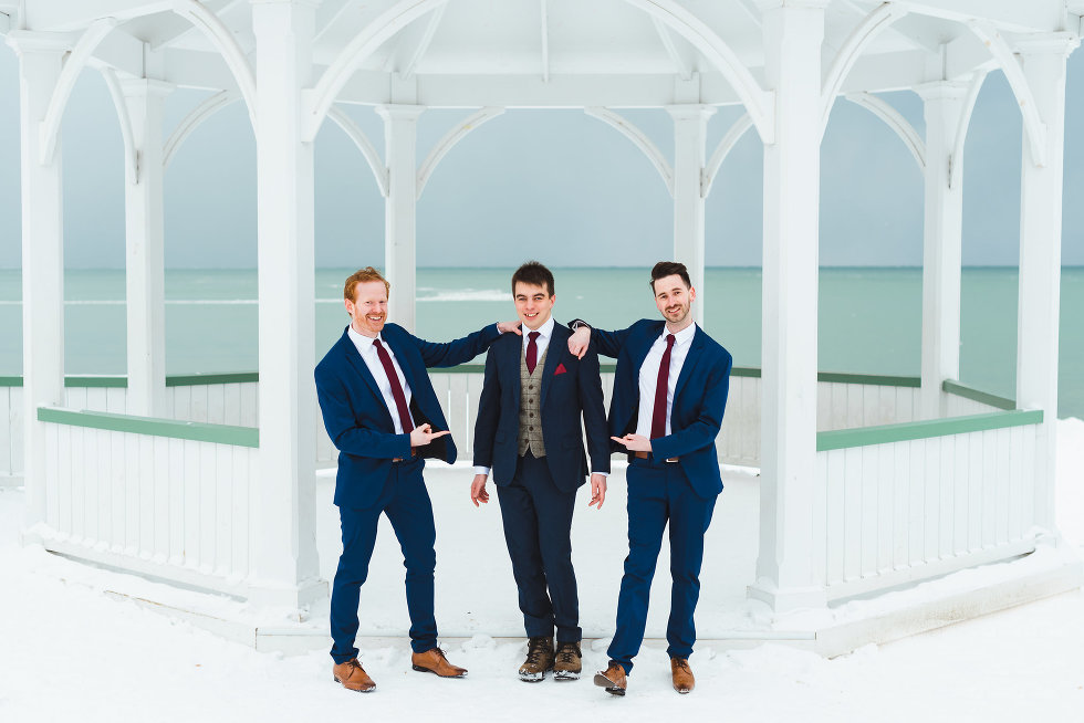 groom stands in front of white gazebo while groomsmen stand on either side of him and point at groom Niagara wedding photography