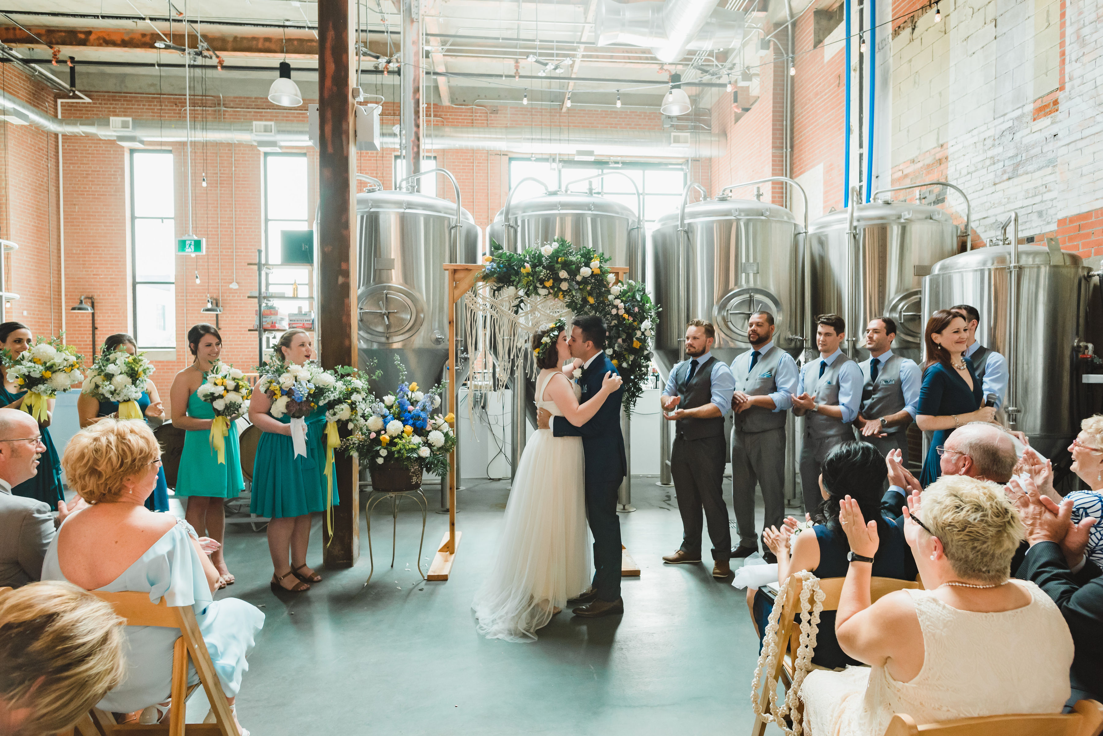bride and groom kiss during wedding ceremony in an old brewery with brewing tanks behind the alter Toronto Junction Craft Brewing