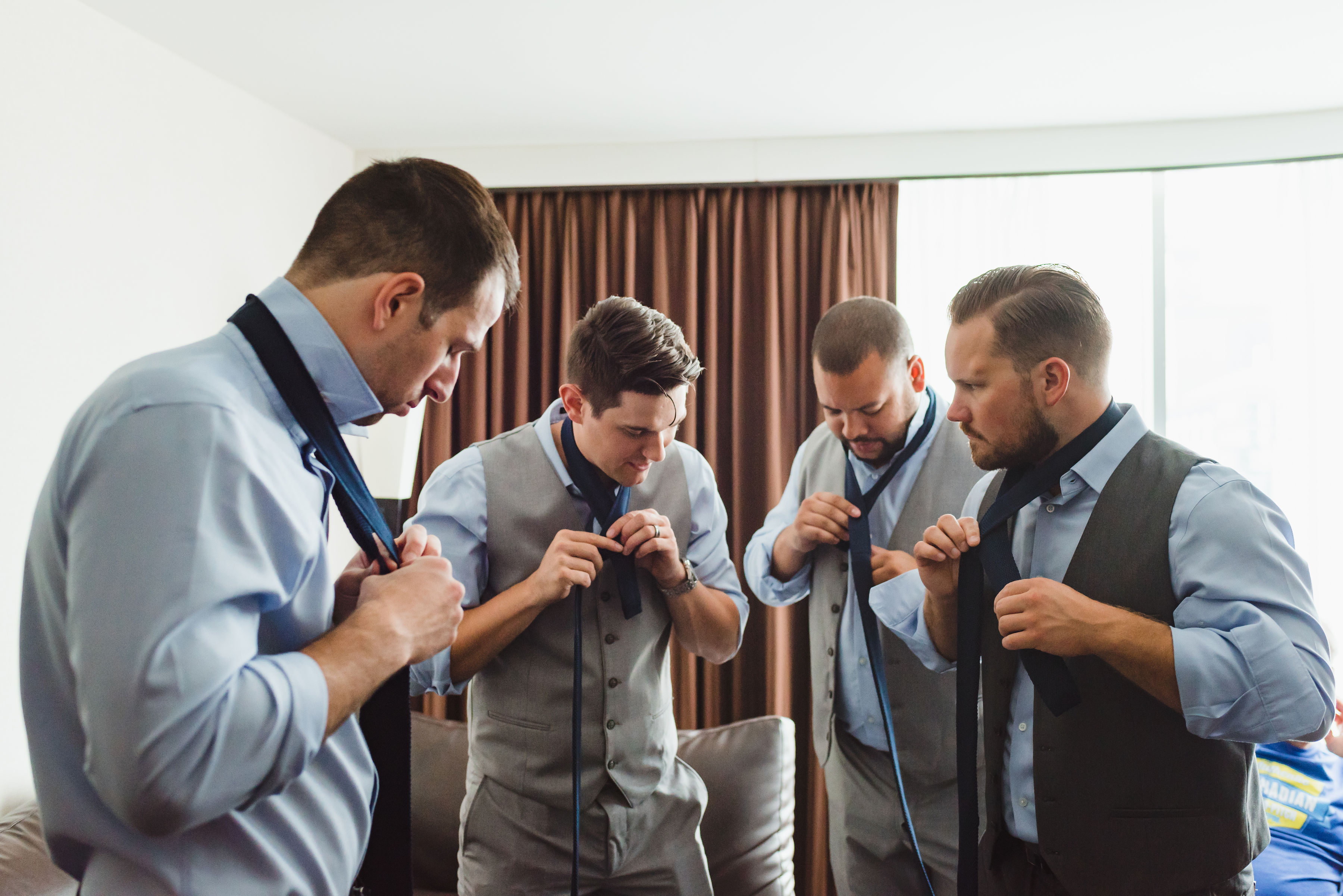 groomsmen in blue dress shirts and grey vests and pants stand in a circle as they all do up their neck ties Toronto Junction Craft Brewing wedding
