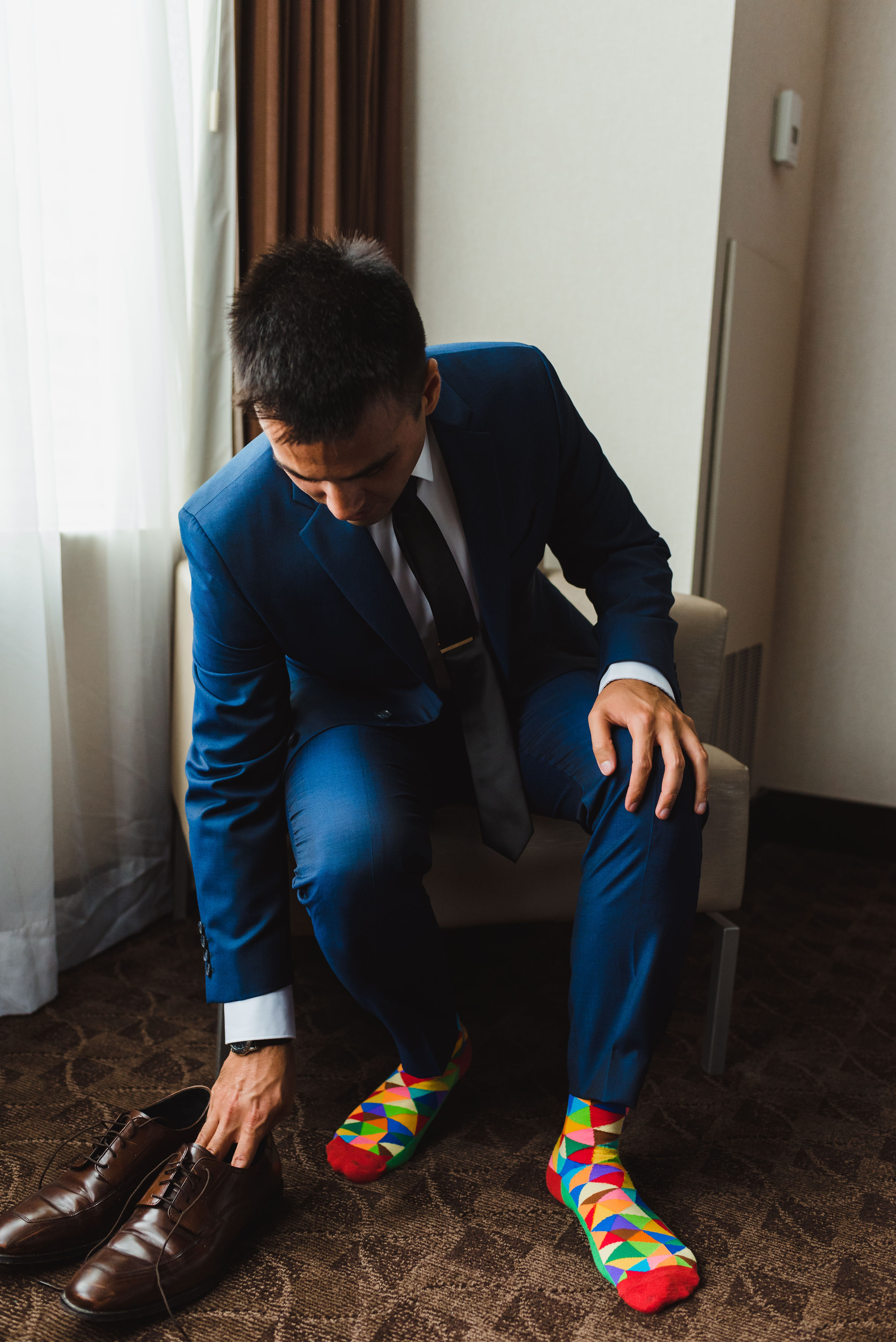 groom in blue suit and multi-coloured socks sitting in a chair about to put on brown dress shoes Toronto brewery wedding photography