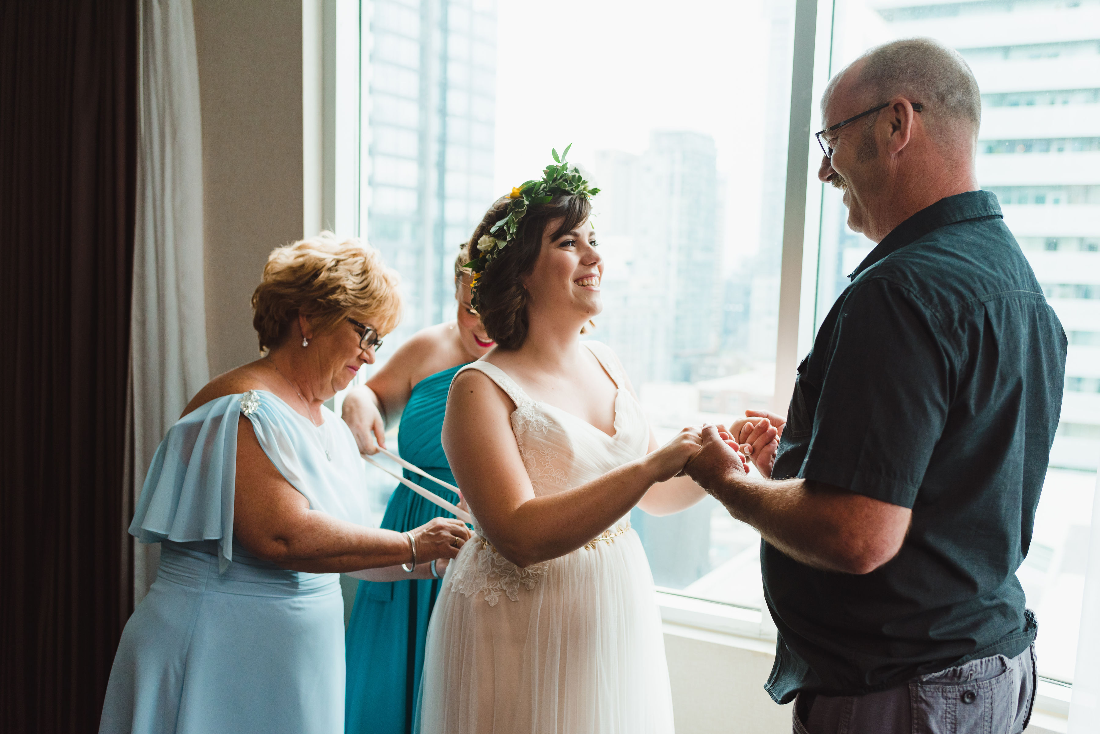 bride having her wedding dress tied up in the back by bridesmaid and mother while she holds her fathers hands in front of her Toronto wedding photography