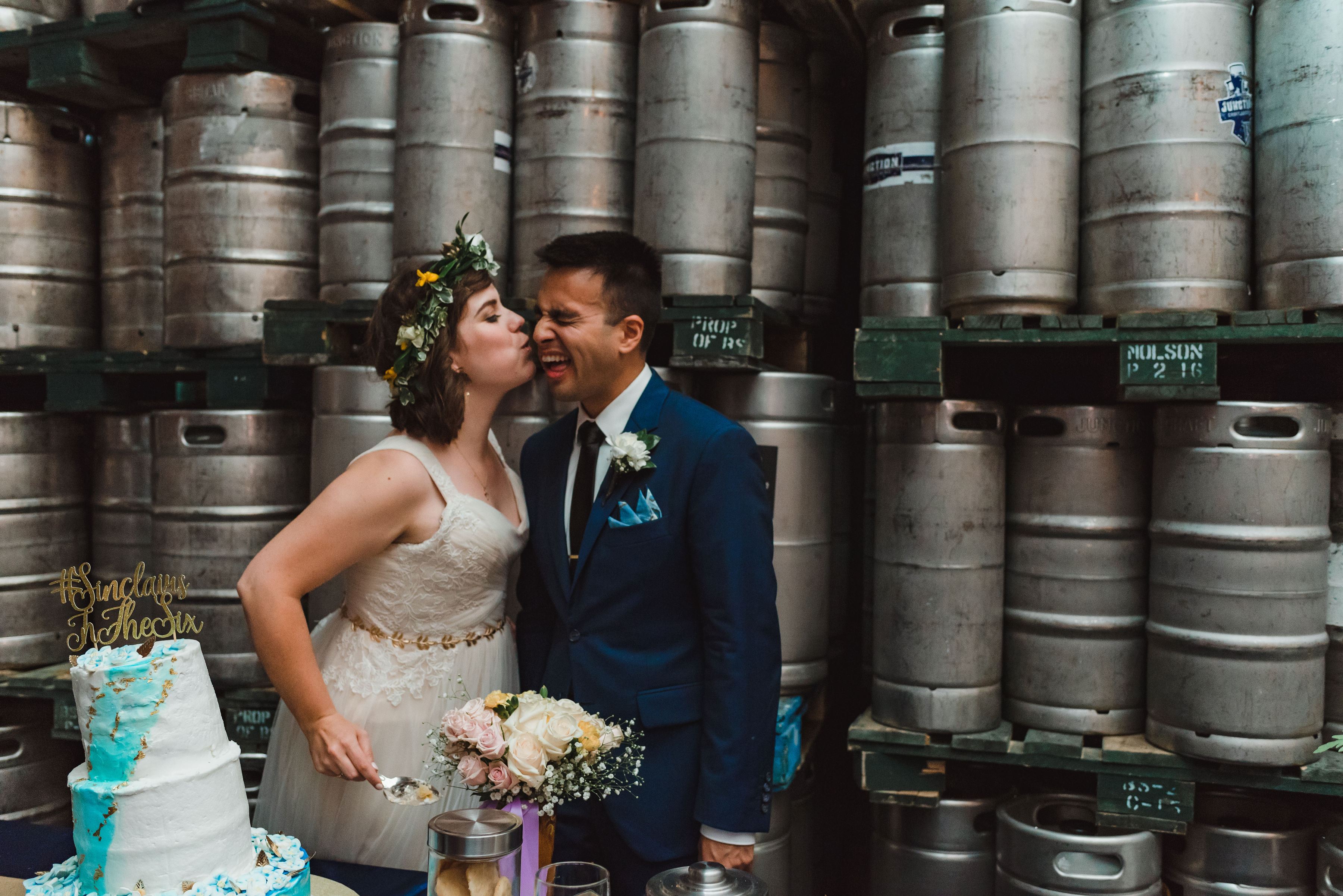 bride kissing groom on his cheek while she cuts the wedding cake with kegs of beer stacked behind them Junction Craft Brewing wedding photography