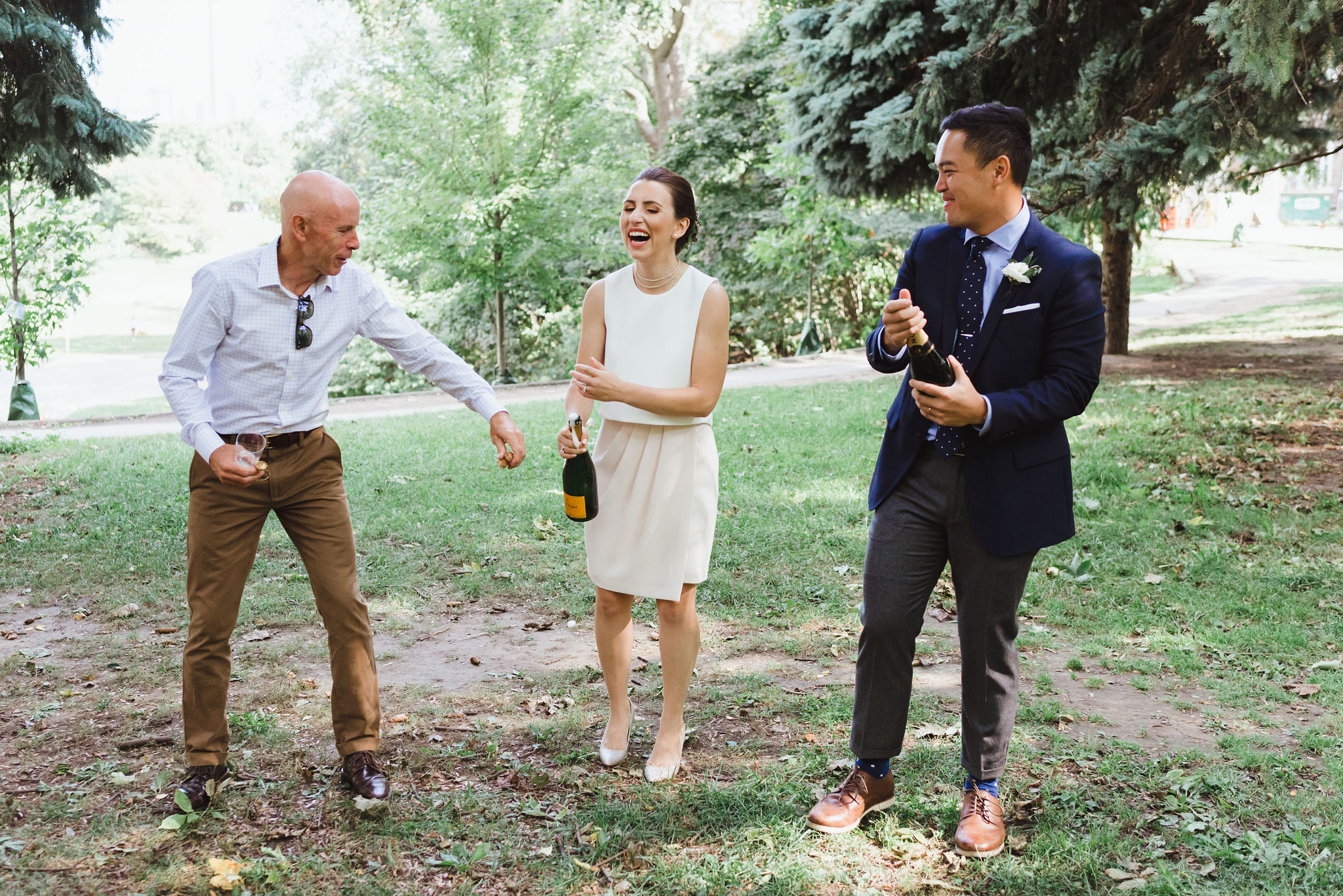 bride and groom opening champagne with her dad reaching over in Trinity Bellwoods Park Toronto wedding ceremony