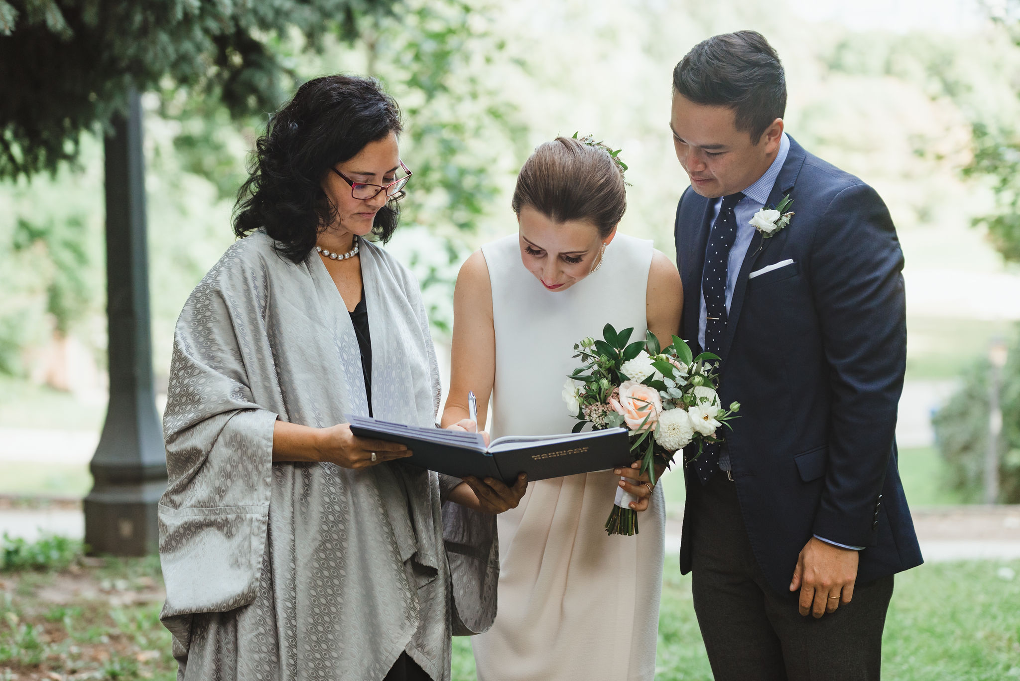 bride signing wedding certificate as groom and officiant look on Trinity Bellwoods Park Toronto