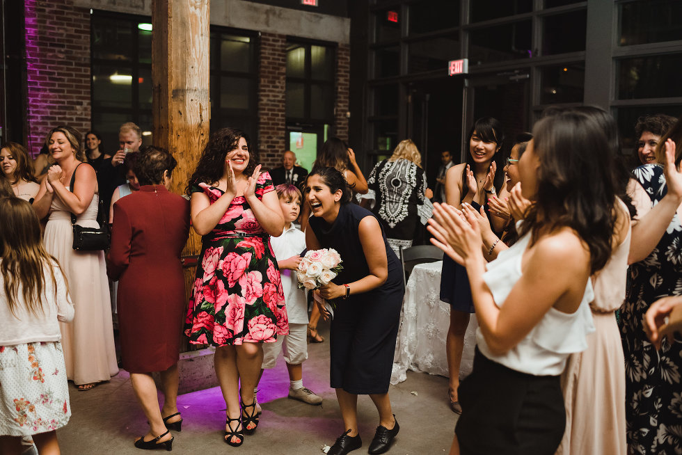 woman laughing as she caught the wedding bouquet as other wedding guests clap around her Steam Whistle Brewing Toronto wedding photography 