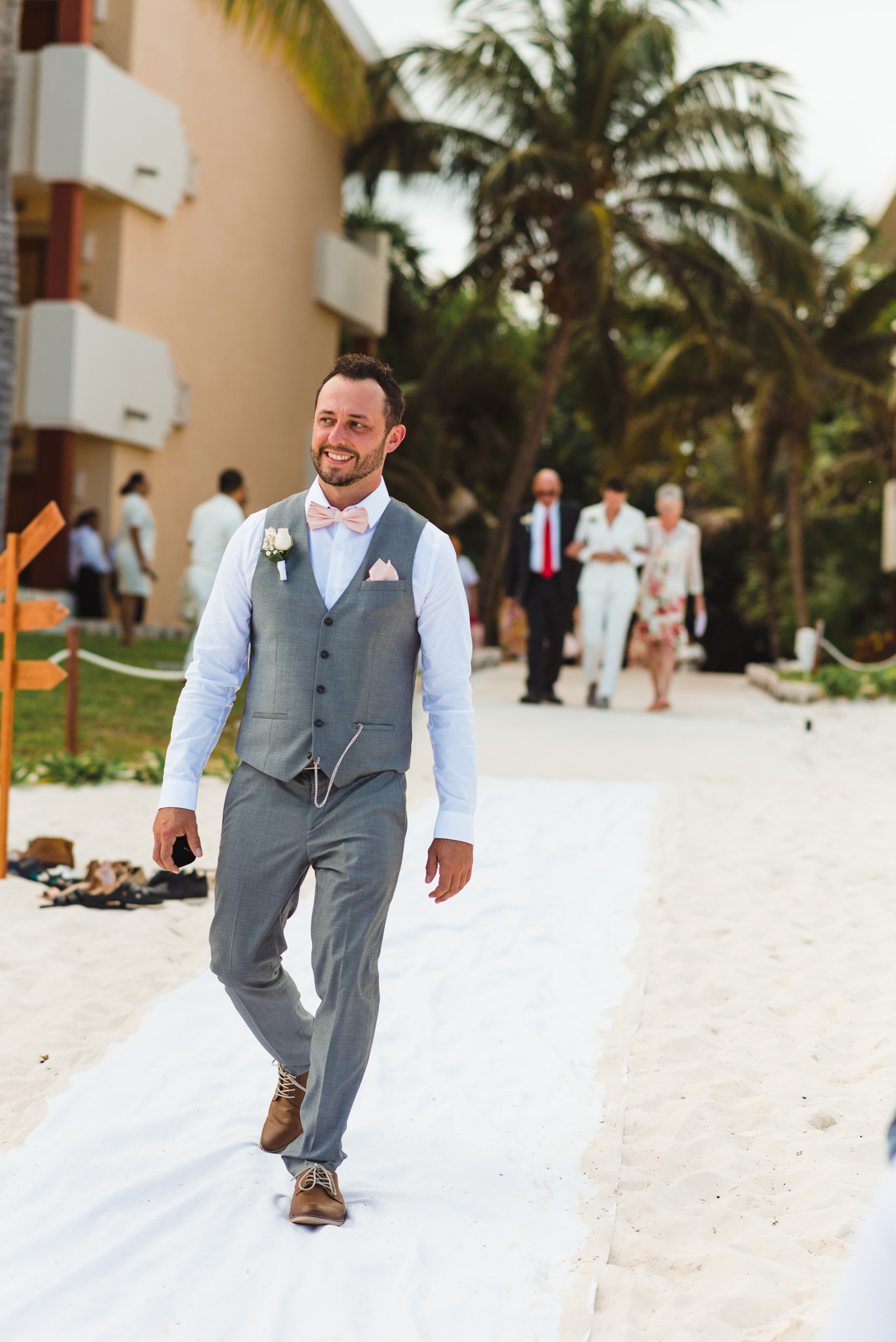 man in grey vest suit and pink bowtie walking into beach wedding ceremony at Now Sapphire Resort in Mexico 