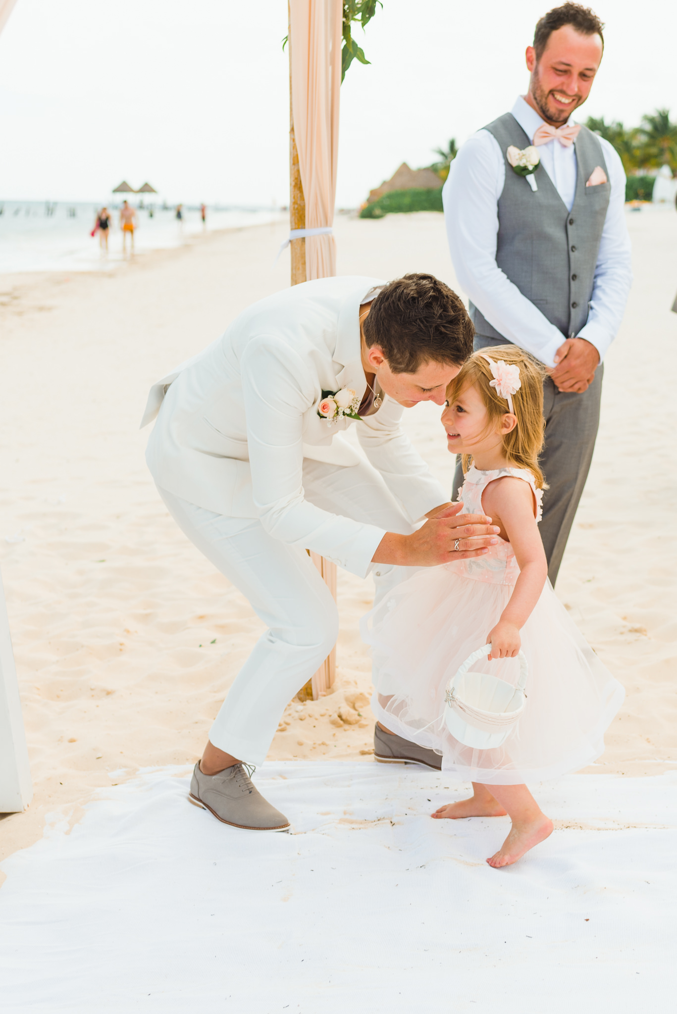 bride crouching down to greet a young girl at the wedding alter as a groomsman watches with a smile during beach ceremony at Now Sapphire Resort in Mexico