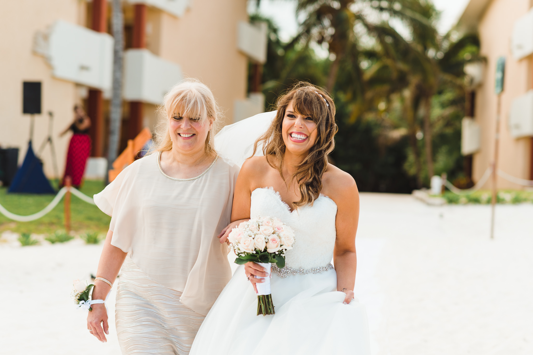 bride smiling and holding bouquet as she's being walked down the aisle by her mother at beach wedding ceremony at Now Sapphire Resort in Mexico