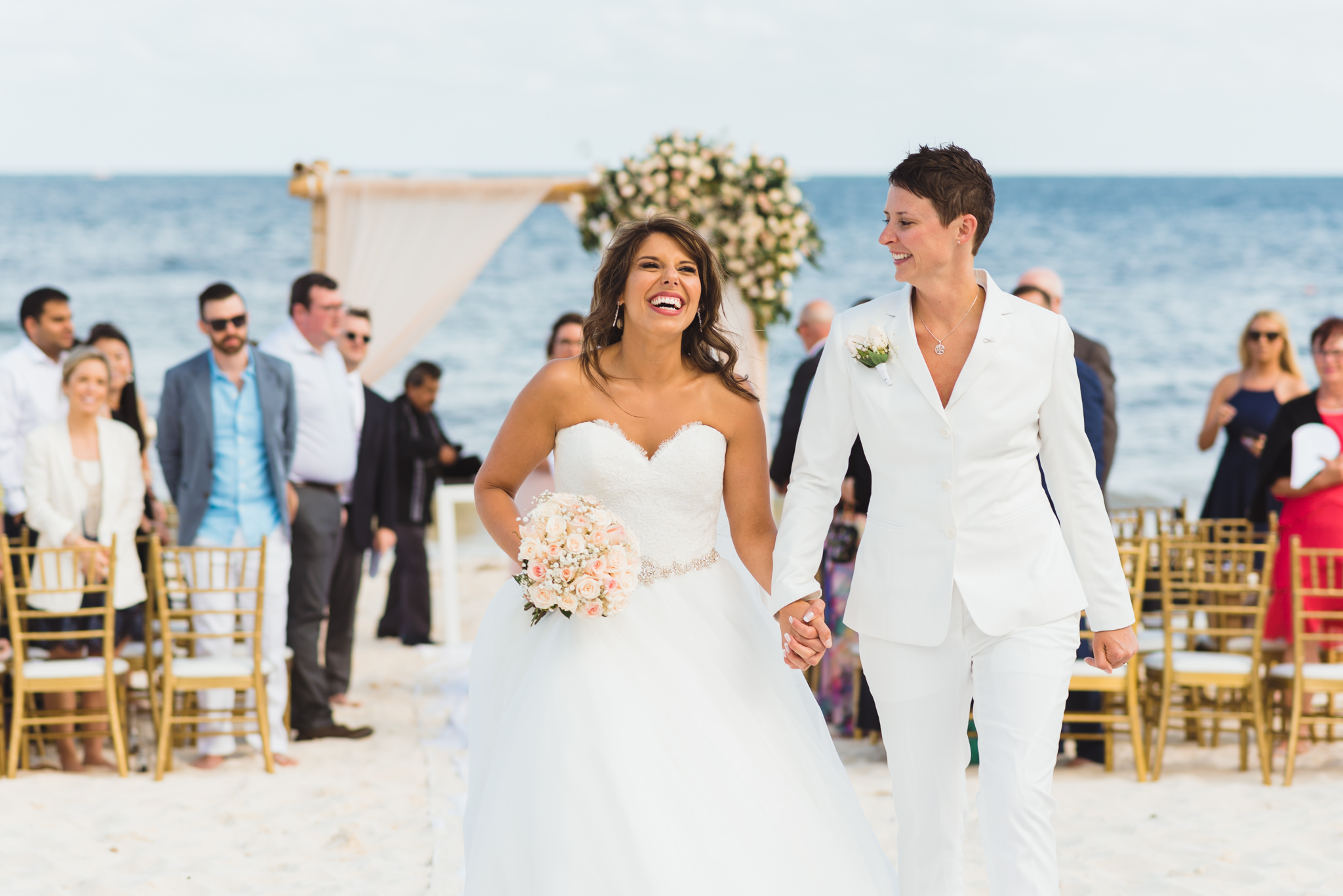 brides smiling and holding hands as they exit beach ceremony in front of ocean at Now Sapphire Resort in Mexico