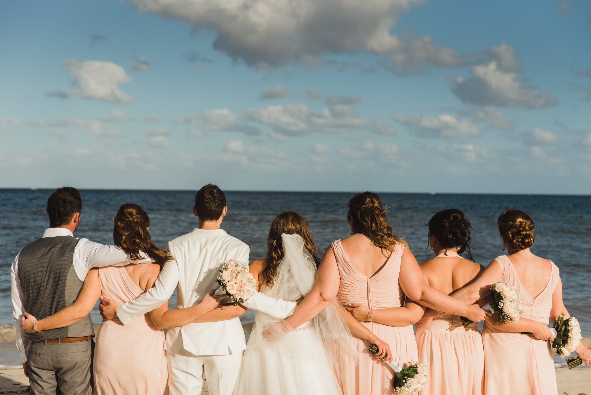 brides and their wedding party facing the ocean with arms around each other on the beach at the Now Sapphire Resort in Mexico how to be really relaxed during your wedding