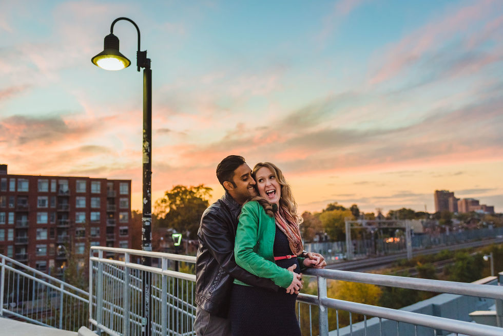 pregnant woman smiling as her partner hugs her from behind on train platform in the Junction, Toronto maternity photos