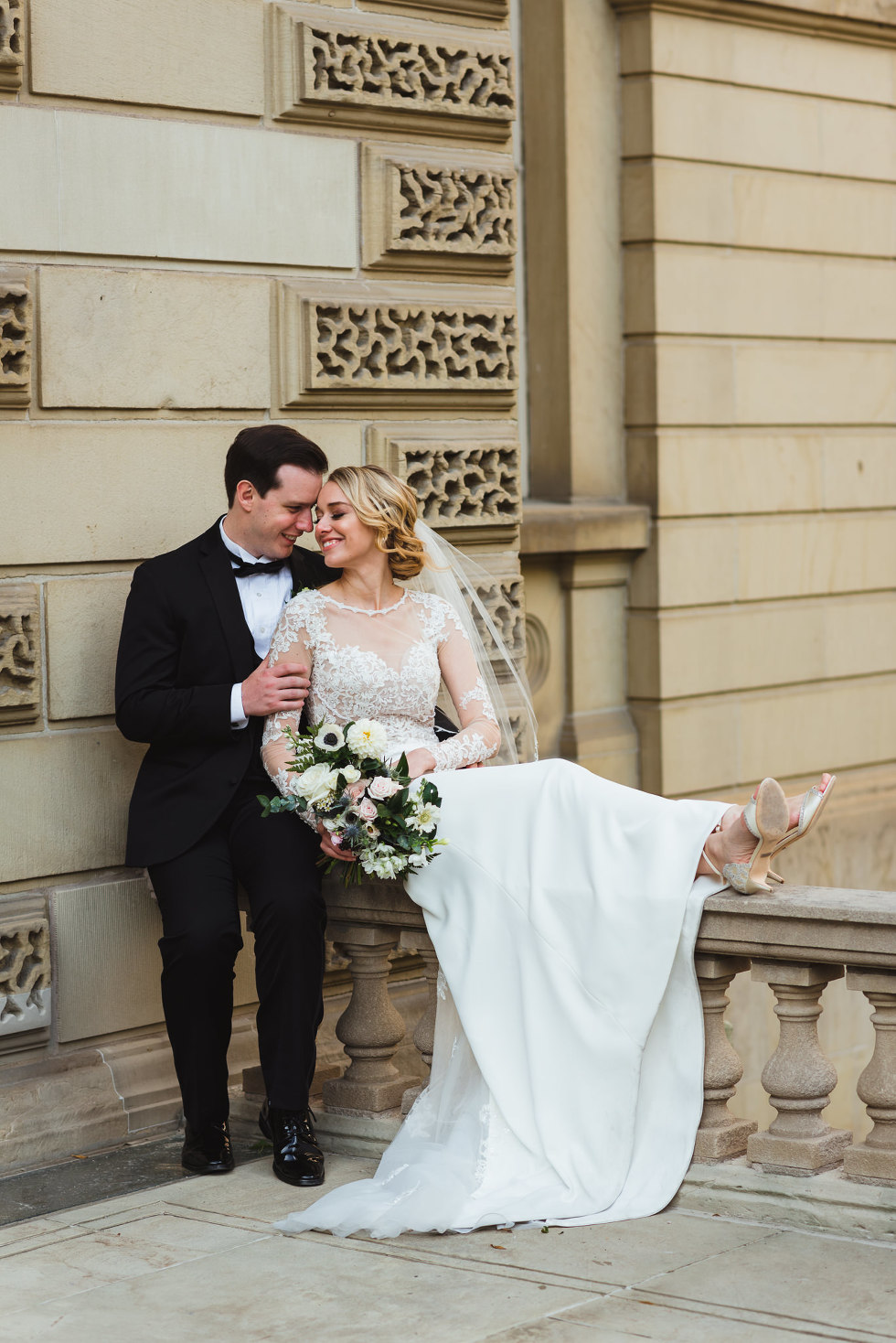 groom standing next to smiling bride as she sits along the railing of an old iconic building in downtown Toronto