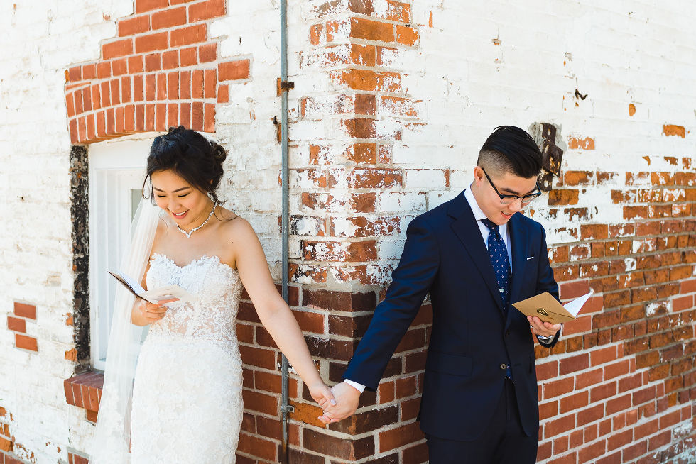 bride and groom wearing a navy suit are holding hands not looking at each other on their wedding day in front of a red and white brick wall reading cards to eac hother at Fantasy Farms in Toronto Ontario