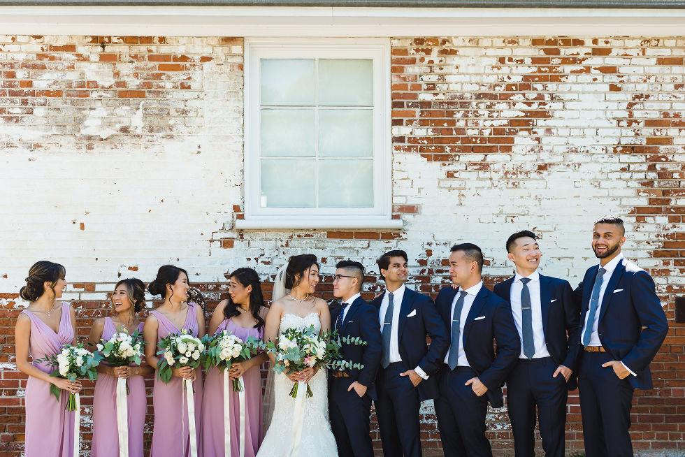 bridesmaids in purple dresses holding green and white floral bouquets with bridal party standing in front of red and white painted brick wall atFantasy Farms in Toronto Ontario