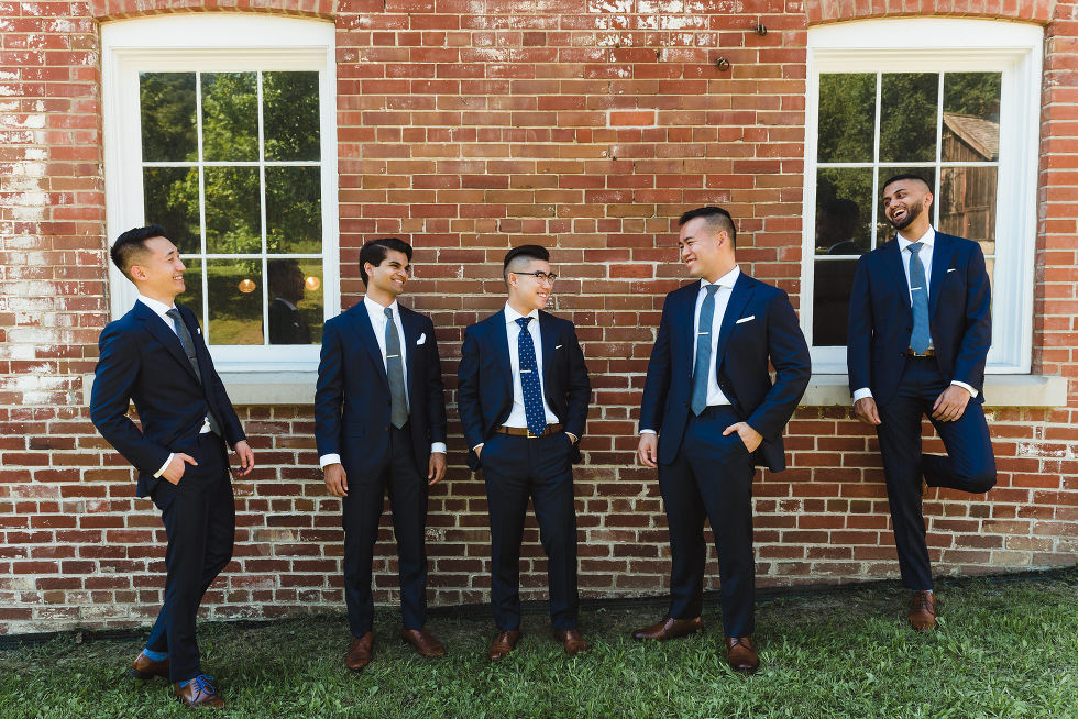 grooms men in navy blue suits laughing in front of old windows and red brick wall at Fantasy Farms in Toronto Ontario