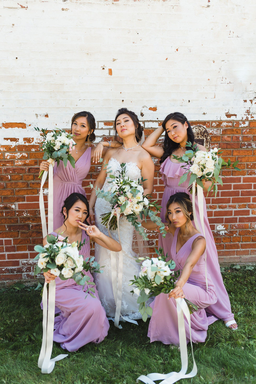 bridesmaid holding green floral leaf and white flower bouquets in lavender coloured dresses in front of white and red brick wall at Fantasy Farms in Toronto Ontario