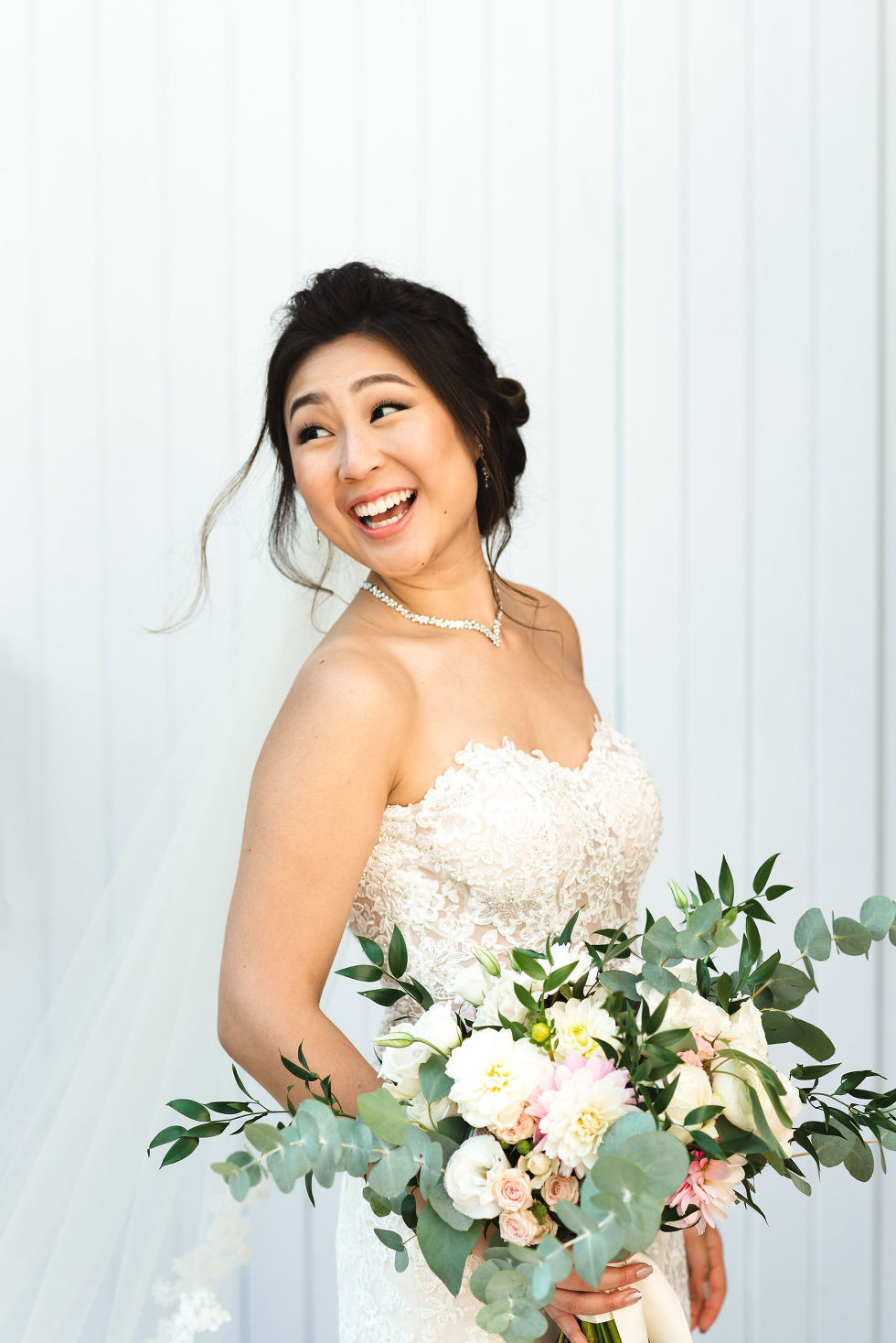 bride smiling with choker necklace on holding green and white flower bouquet in front of white barn doors at Fantasy Farms in Toronto Ontario