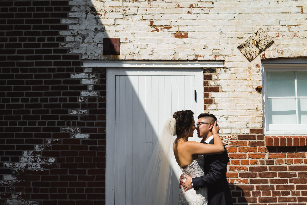bride kissing groom in front of white barn doors and red brick wall with cool sun shadows going across them at Fantasy Farms in Toronto Ontario
