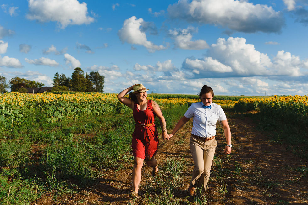 2 women walking hand in hand into a stunning sunflower field as one woman puts a hand over her fedora on her head Toronto engagement photos