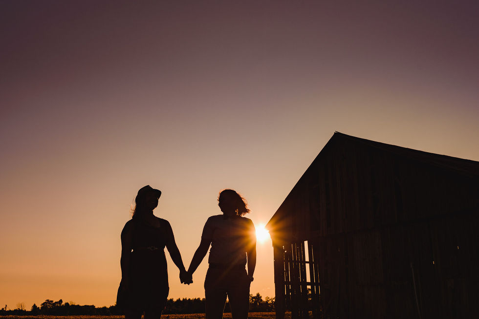 engaged couple holding hands in front of a barn with the sun setting behind them stunning sunflower engagement photos