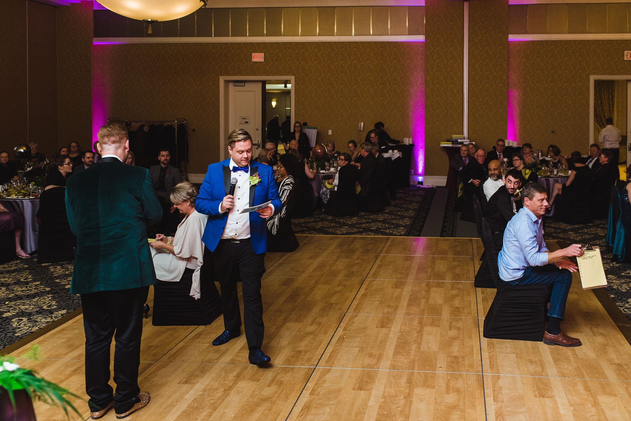 grooms reading game instructions into their microphones for their seated guests that are scribbling away on clipboards during a fun wedding at the Hilton Fallsview in Niagara Falls