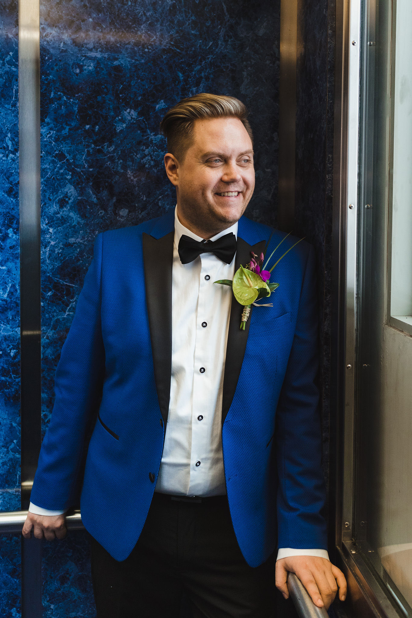 groom in a bright blue suit stands in front of a blue marble wall at the Hilton hotel in Niagara Falls before his wedding