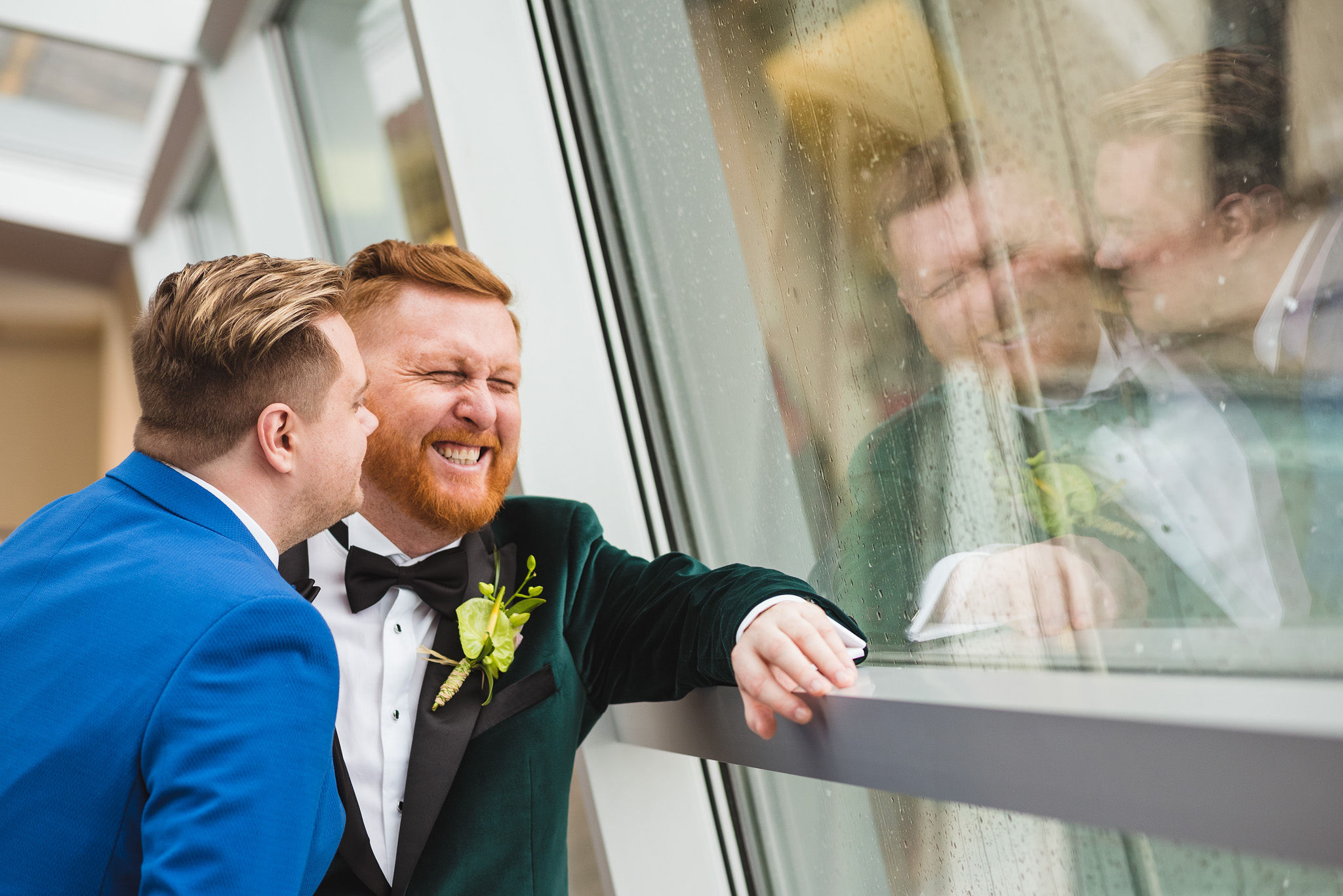 groom in bright blue suit leans into groom in green suit and makes him laugh before their wedding at the Hilton Fallsview in Niagara Falls 