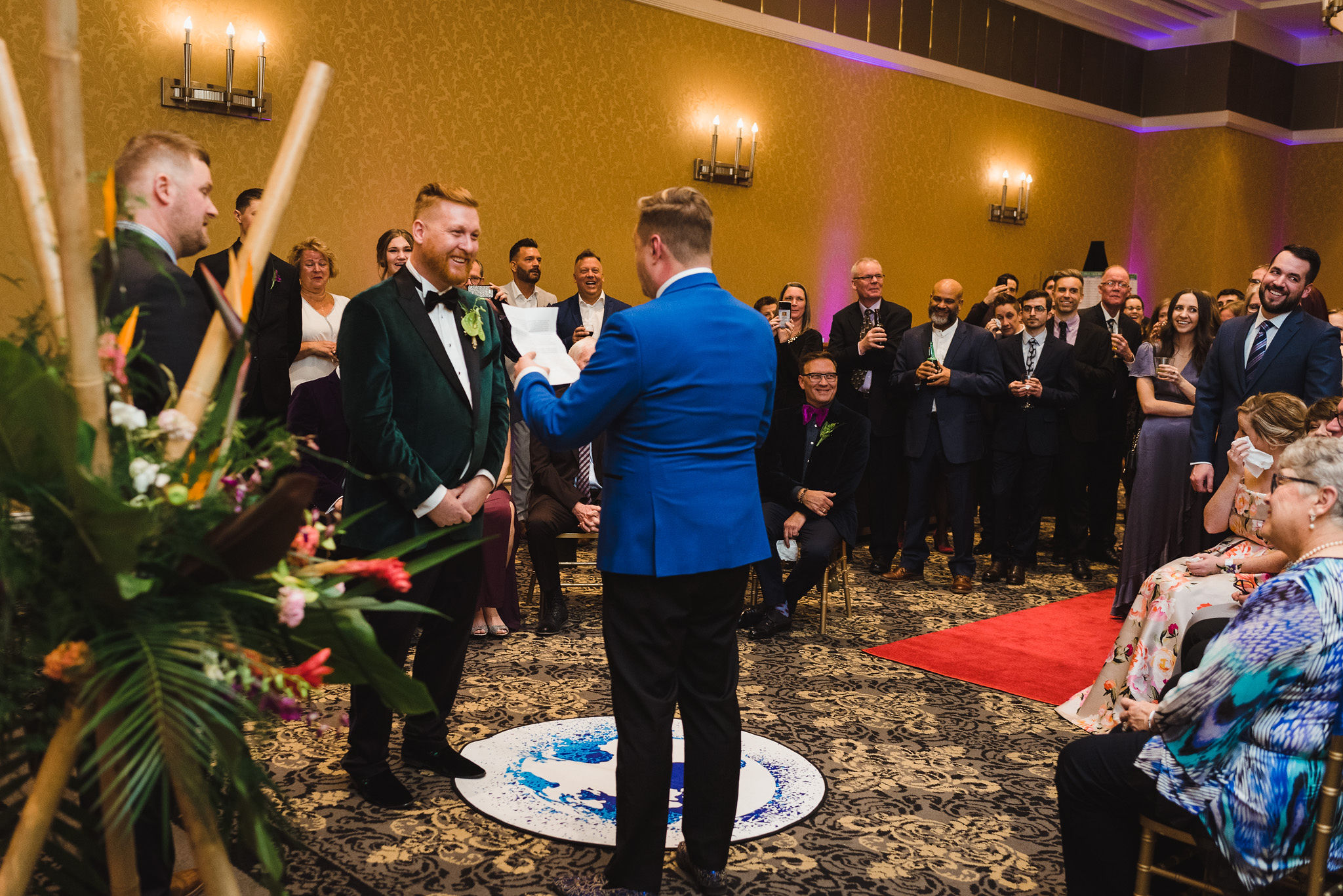 groom smiles and wedding guest look on as the other groom reads his vows aloud during wedding ceremony at the Hilton Fallsview in Niagara Falls