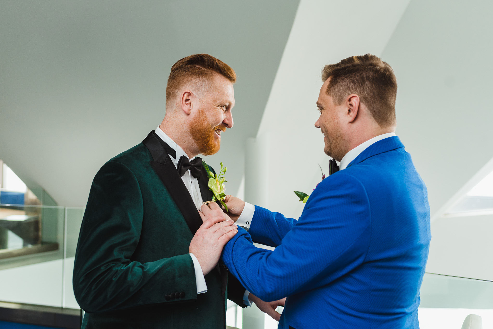 Groom in blue suit placing boutonniere on lapel of groom in green suit before their wedding at the Hilton in Niagara Falls 