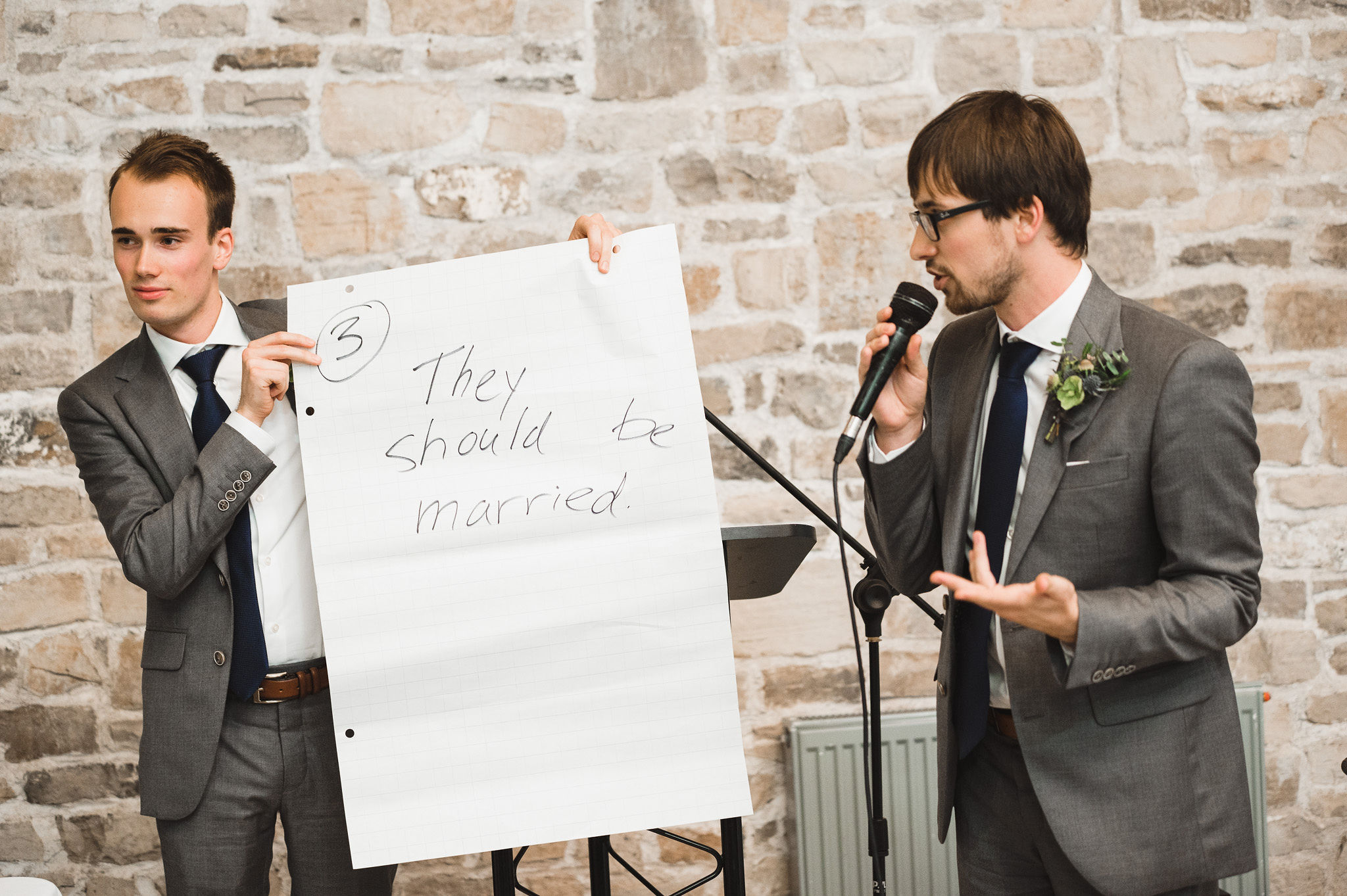 A couple groomsmen holding up a large sheet of paper with "They should be married" written on it as one of them reads into the microphone during a charming southern style wedding at Ruthven National Historic Site