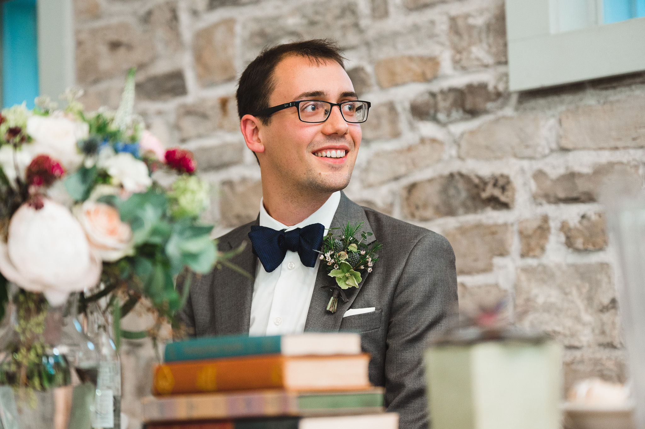 groom smiling at his head table in front of old stone wall with books and flowers on the table in front of him during his wedding at Ruthven National Historic Site
