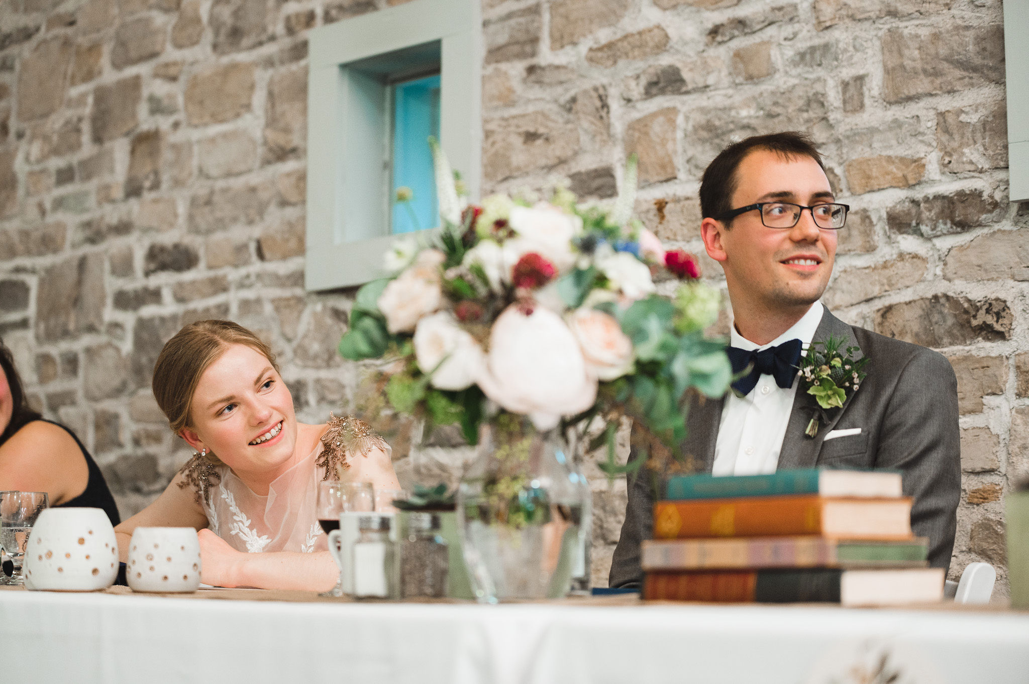 bride and groom smiling at their head table in front of old stone wall with books and flowers on the table in front of them during speeches at their wedding at Ruthven National Historic Site