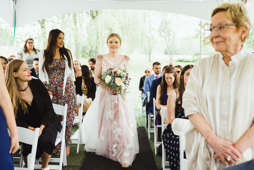 bride holding floral bouquet beginning her walk down the aisle past her onlooking guests during her charming southern style wedding at ruthven national historic site