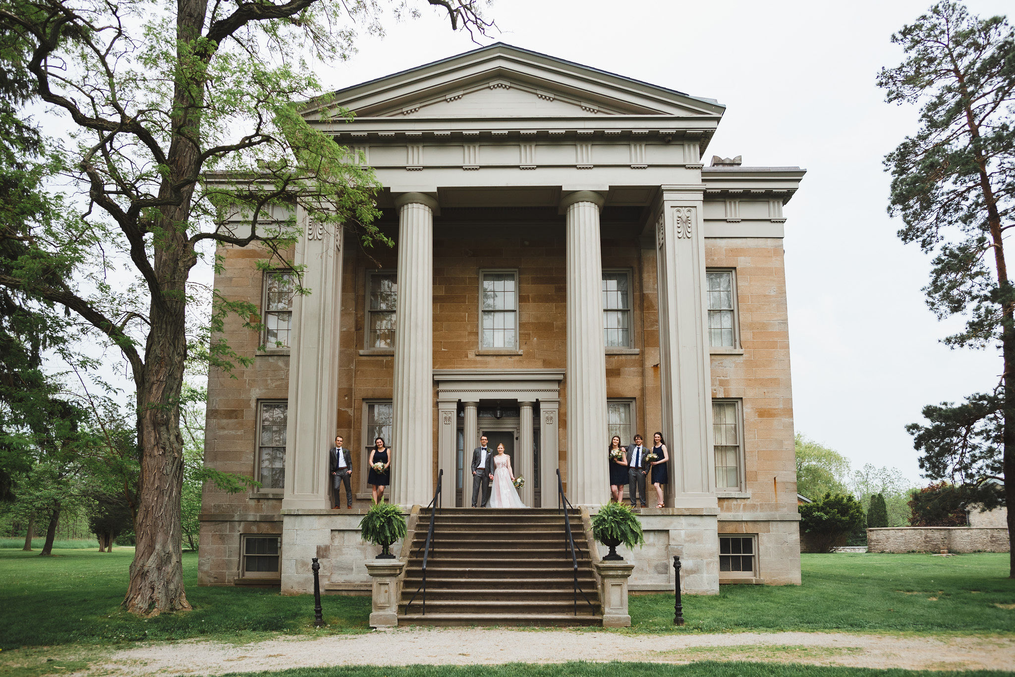 bride and groom standing between their wedding party on stone steps between Greek stone columns of the Ruthven National Historic Site after their charming southern style wedding near Hamilton