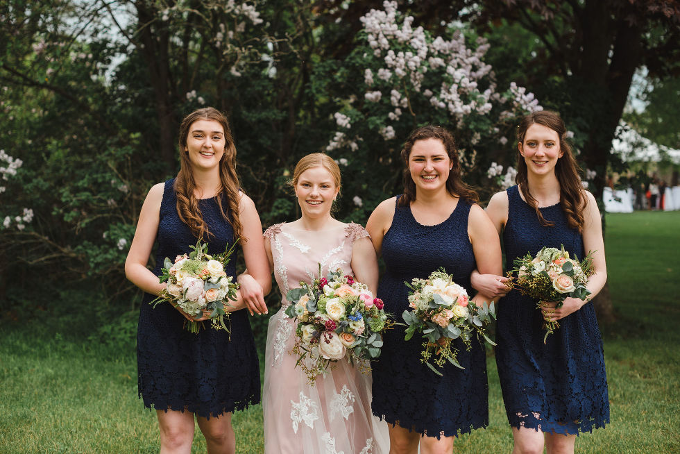 bride standing between her three bridesmaids with arms linked as they all hold floral bouquets at Ruthven National Historic Site during her charming southern style wedding near Hamilton