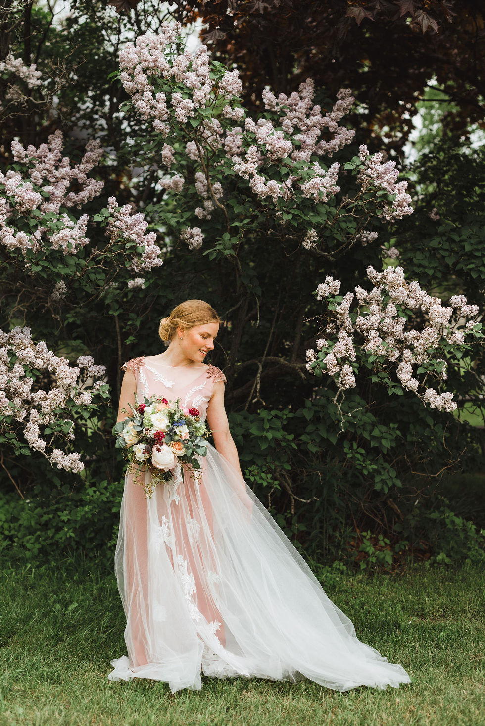 bride standing in front of lilac bushes holding a floral bouquet and looking down at her dress during her charming southern style wedding at Ruthven National Historic Site near Hamilton