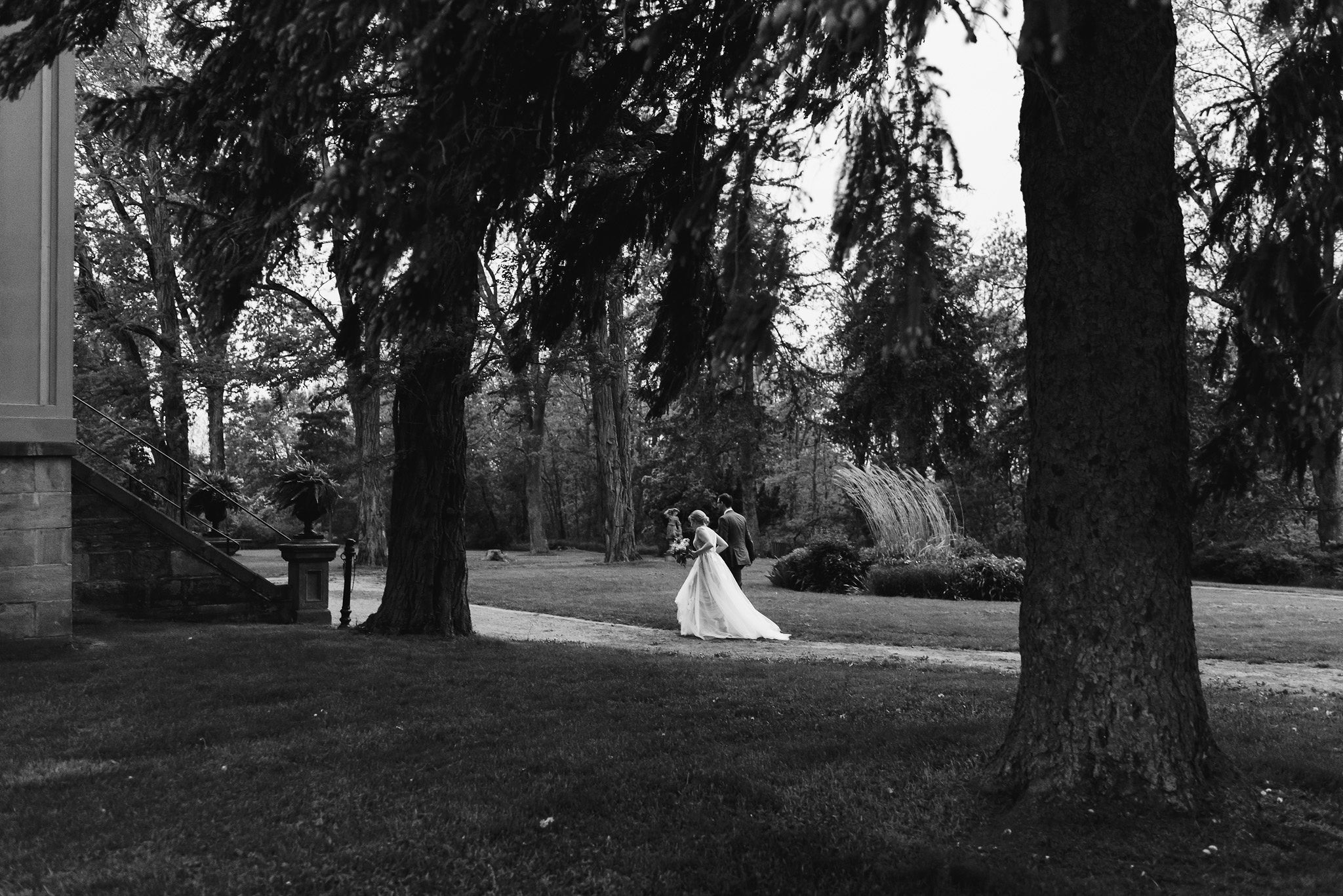 bride and groom walk along a forested path leading to a stone building during their charming southern style wedding at Ruthven National Historic Site near Hamilton
