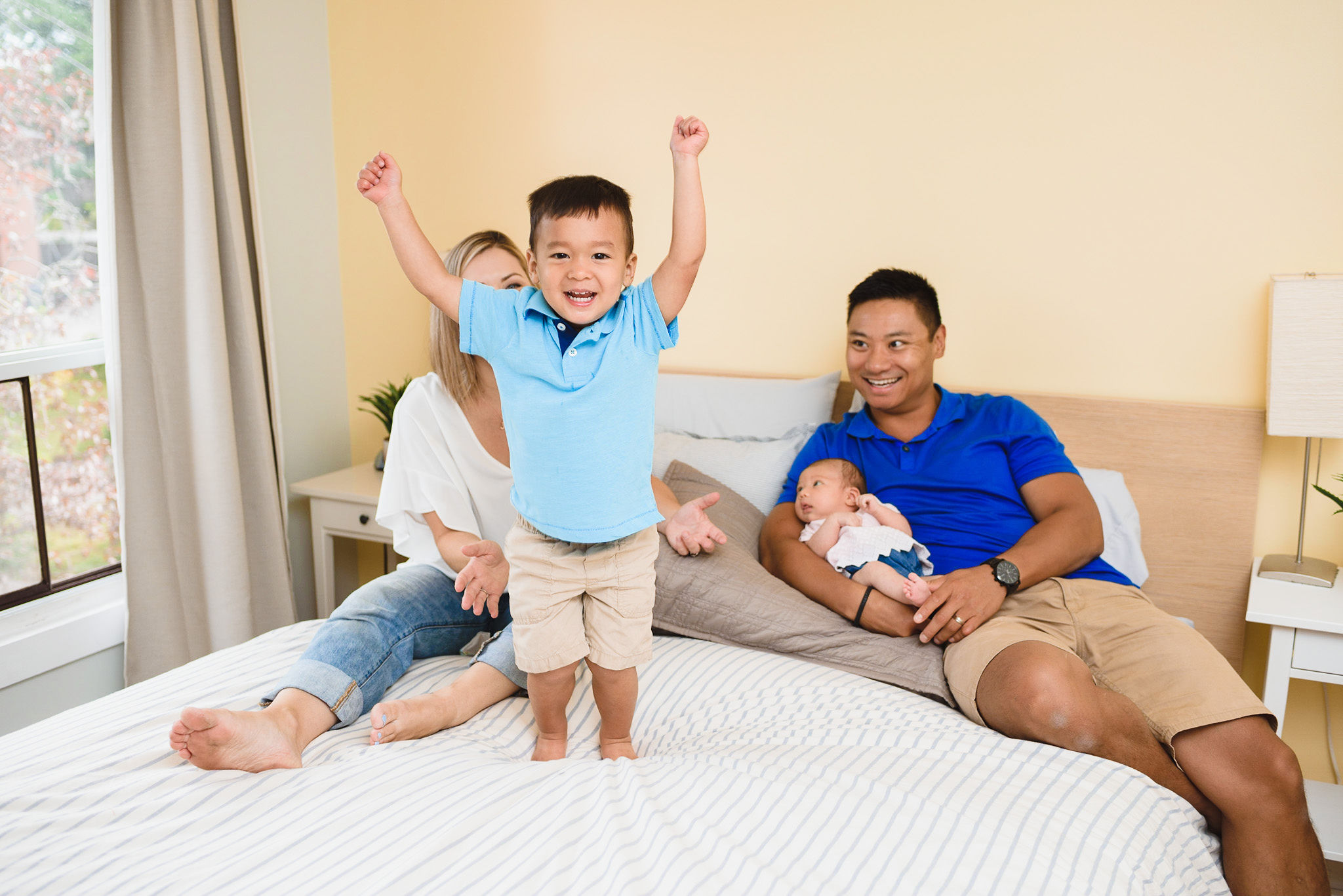 Husband and wife laying down on their bed with a newborn in dads arms and a toddler standing on the bed with his arms in the air Toronto family photography