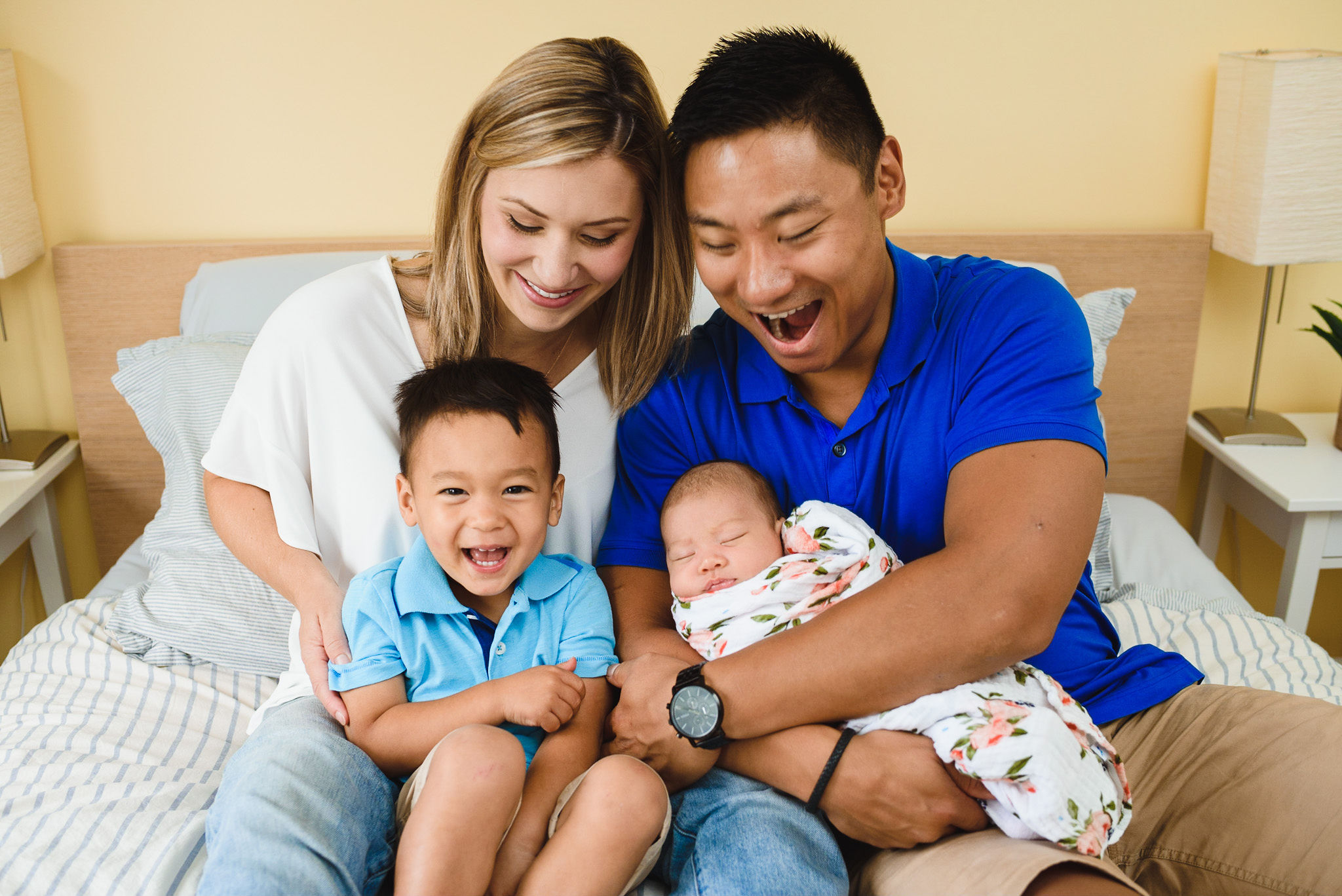 A mother and father laughing while sitting on their bed with their toddler and newborn in their laps Toronto family photos