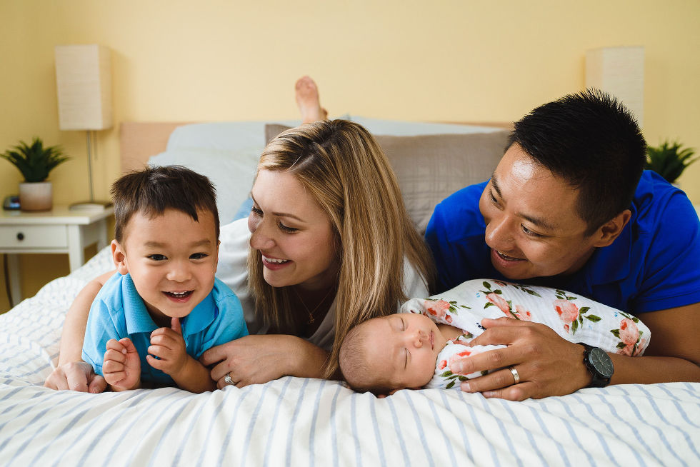A mother, father, and toddler laying on their stomachs on their bed with a swaddled baby laying in front of them Toronto family photography