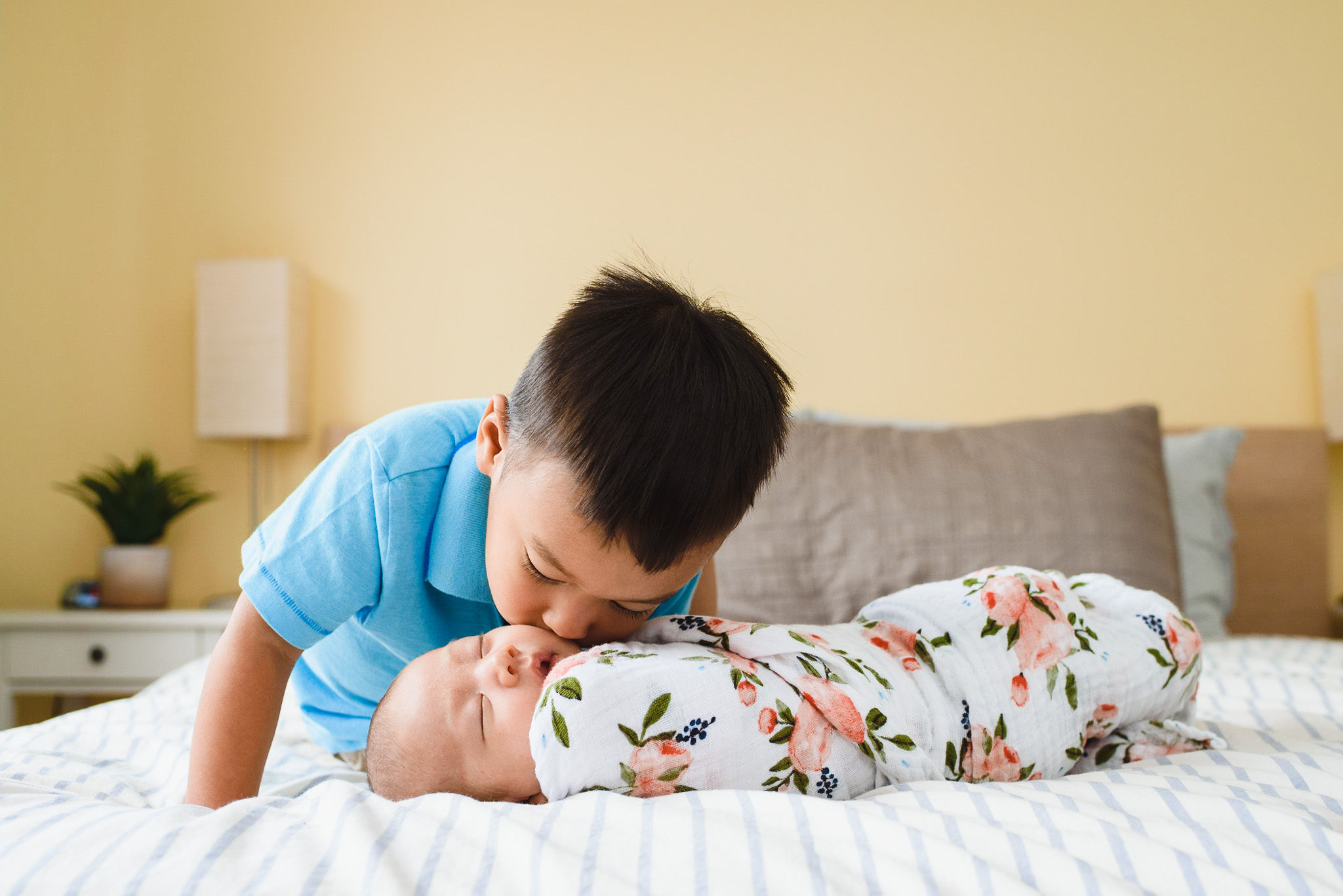 toddler crouching down to kiss his newborn sibling that's swaddled in a floral blanket Toronto family photography