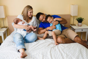 A mother holding her newborn baby as her toddler is wrapped up into the arms of the father sitting next to her on their bed Toronto family photography